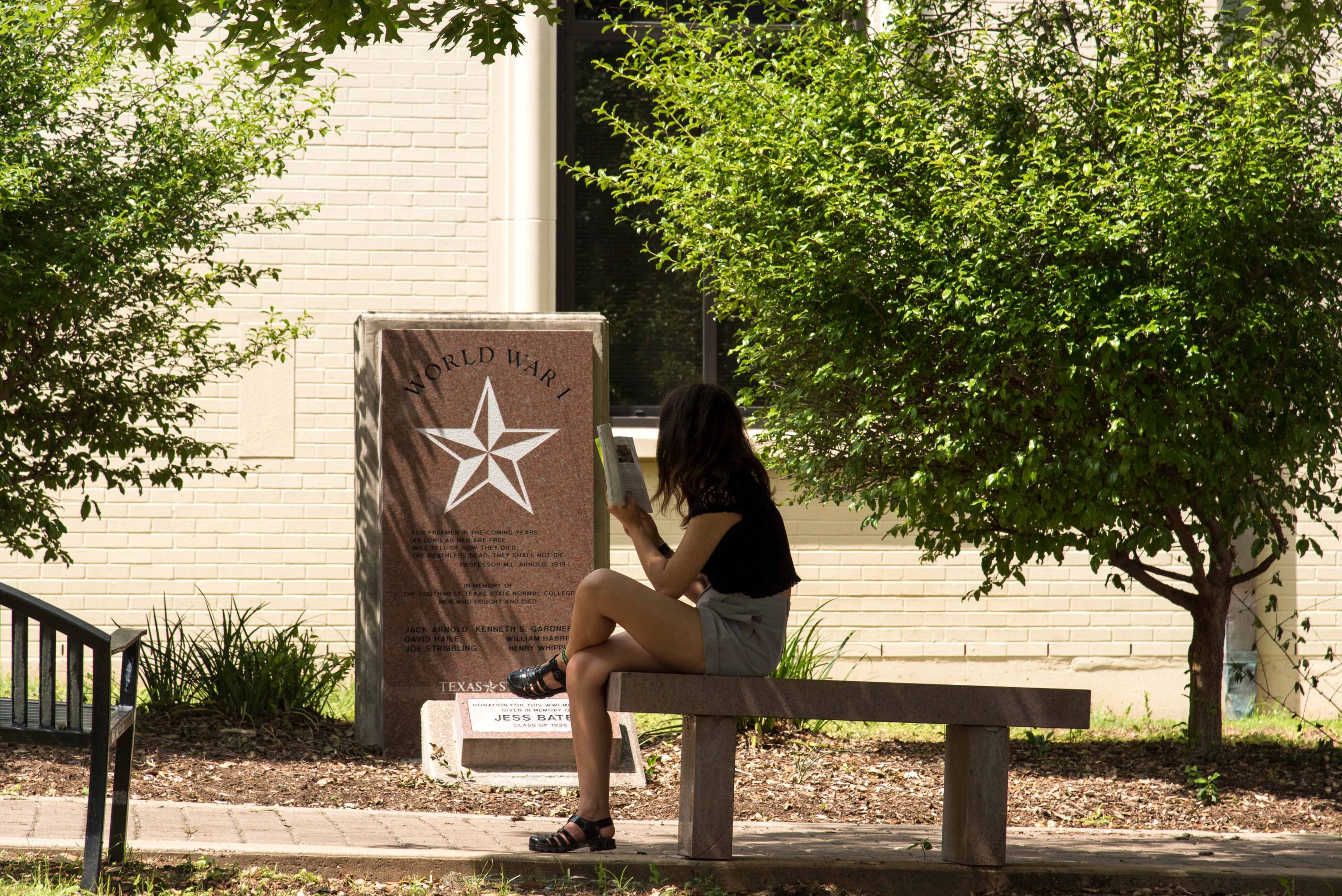 Photograph of the World War 1 monument in the Veterans Memorial Garden at Texas State University's campus today. It is a simple, rectangular slab of red granite bearing a white star and 'World War 1' emblazoned across the top. Beneath it, the names of students who died in the war are engraved. A young woman sits in the shade of the trees reading a book on a bench across from the monument.