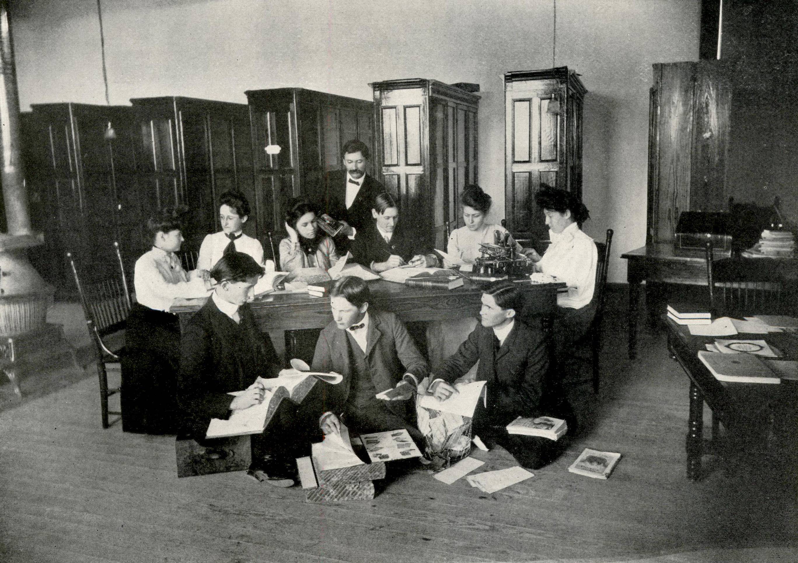 Aged black and white photo, showing a group of students working in a library. 