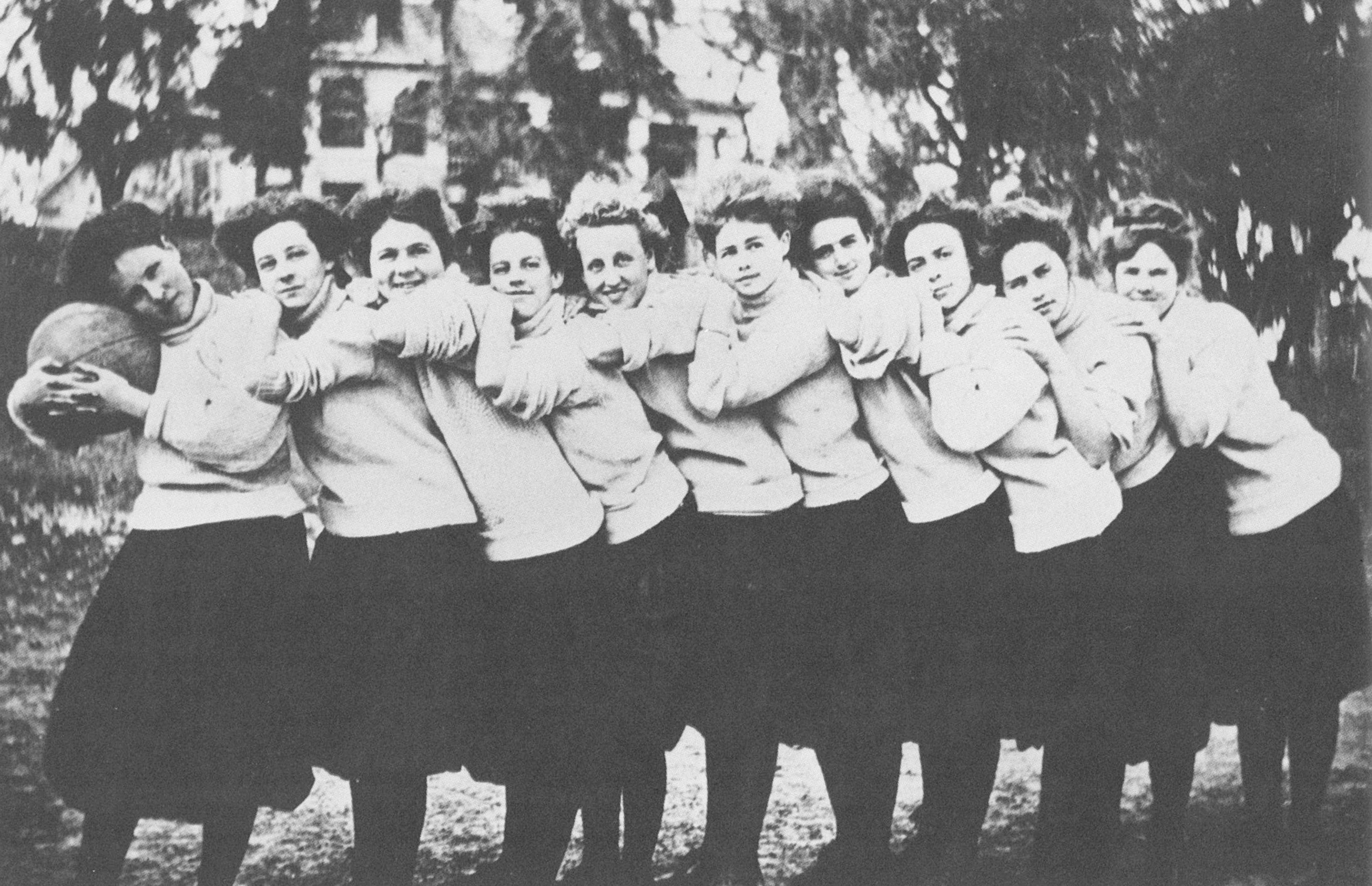 Aged casual photo of the women's basketball team. The young women are all wearing their game uniforms, which consist of a bulky sweater and dark mid-calf length skirts. They're standing in profile, in a row, each girl leaning on the girl in front of them, hugging her shoulders. The first woman in the row leans on a basketball.