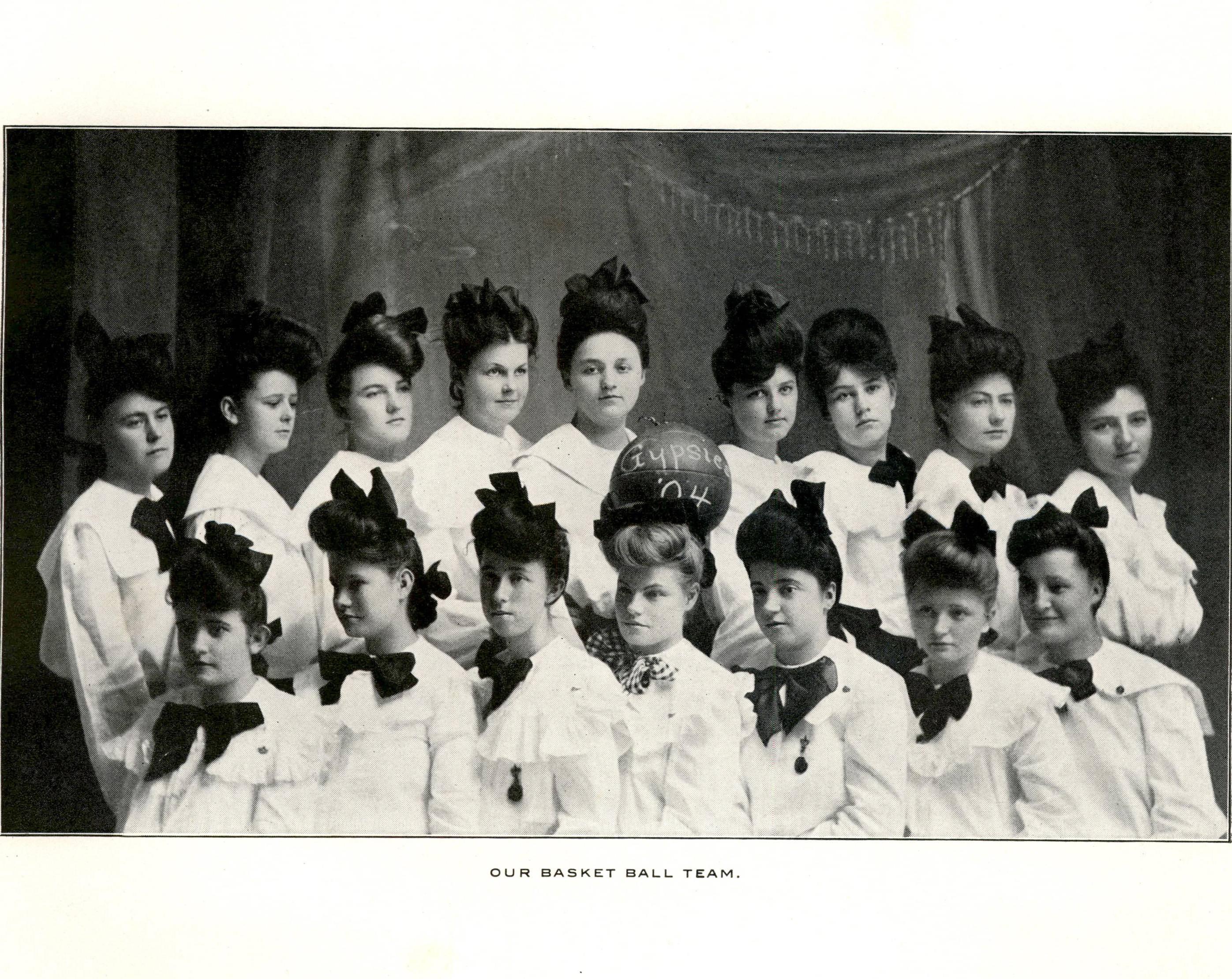 Aged team photo of the women's basketball team. They're seated in two rows, and wearing matching white shirts and large dark hair bows. One woman in the center of the top row holds a basketball.