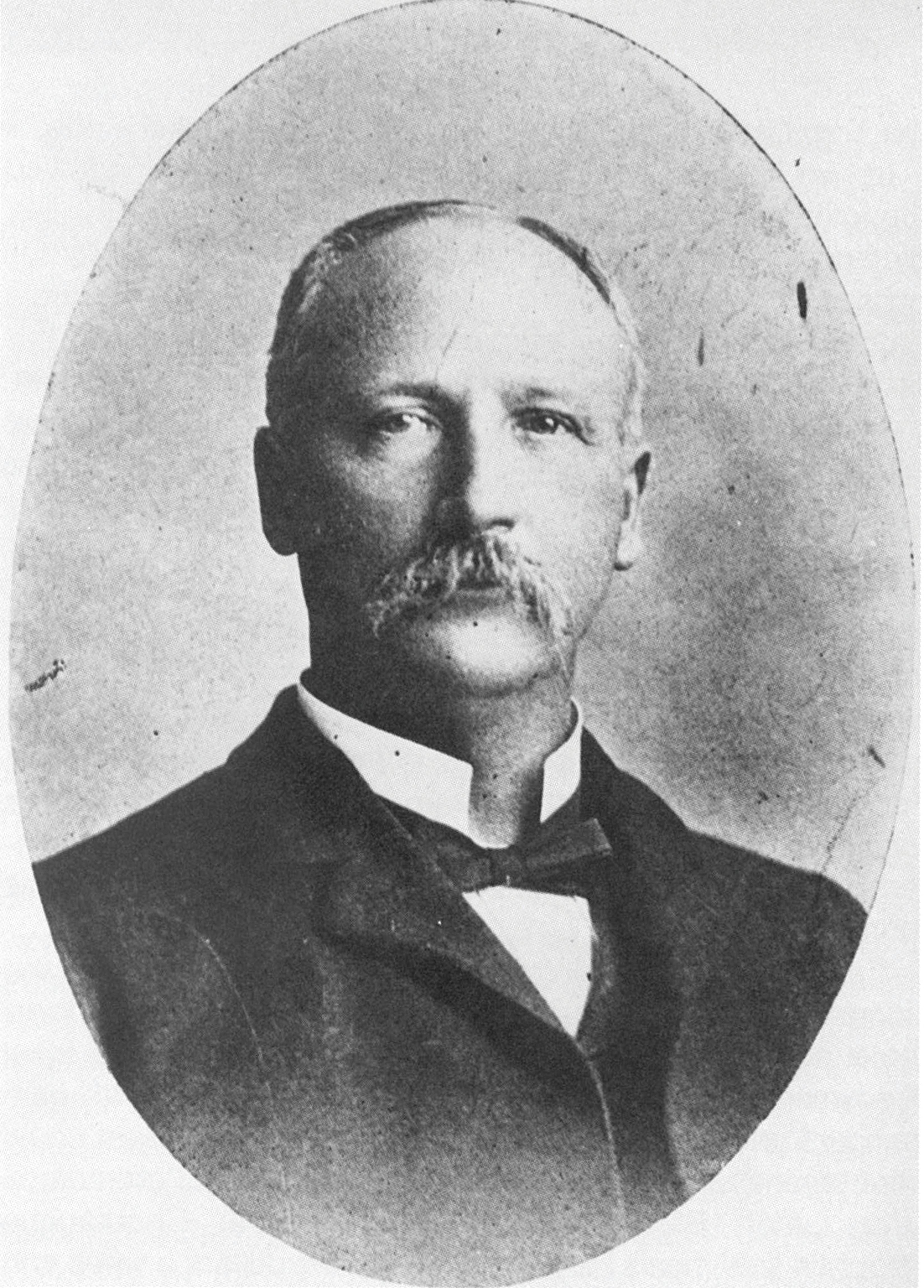 Aged black and white portrait of Thomas Harris. Harris is a white man and appears to be in his 40s, he has a large, bushy mustache, and receding light-colored hair, trimmed tidily. He's dressed in a typical Edwardian fashion, wearing a dark jacket and bowtie, with a stiff upright white collar.