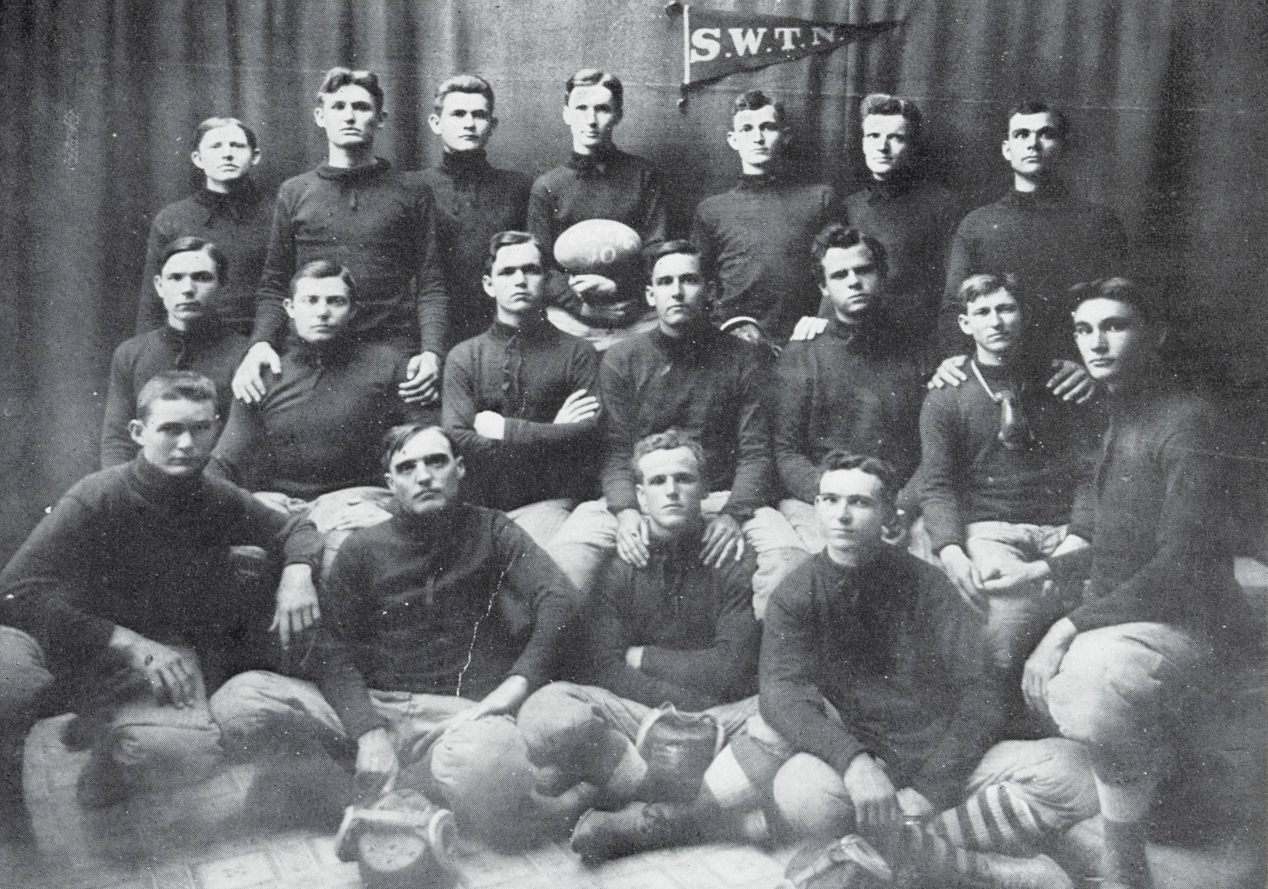 Black and white yearbook photo of the first football team. The young men are posed in front of a dark curtain in three rows, the first row kneels, the second is seated, and the third stands. They're wearing game uniforms which consist of a dark, long-sleeved turtleneck shirt, and light pants tucked into striped socks. A man in the center of the third row holds a football, and a pennant reading 'S.W.T.N.' is pinned to the curtain behind them.