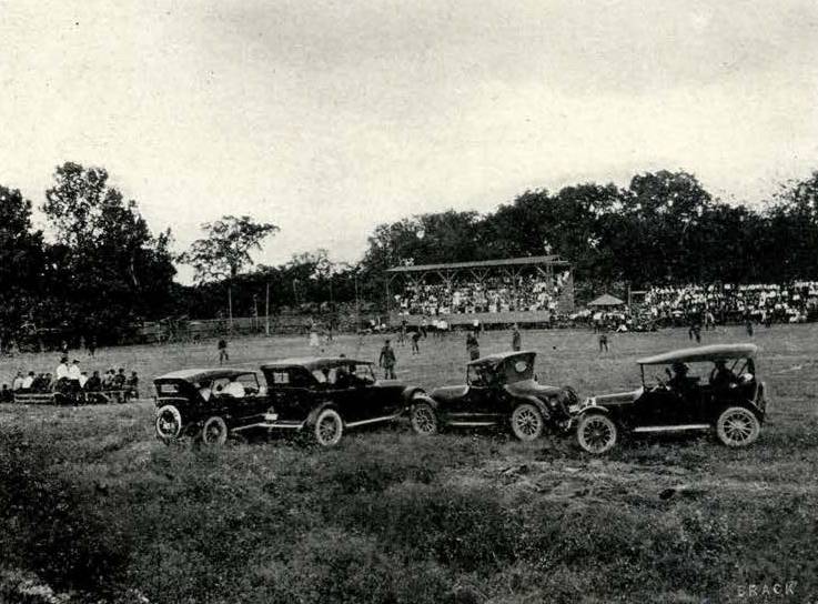 Aged black and white yearbook photo of Evans Field the day of the first homecoming game. The weather is clear, and in the foreground, several old cars are parked in the grass as attendees watch the game from a few benches set off to the side. Past the cars, the football players are scattered across the field mid-game. In the background, a small stand of covered bleachers has been erected, as well as a few small tents and more uncovered bleachers, and are all filled with people watching the game.  