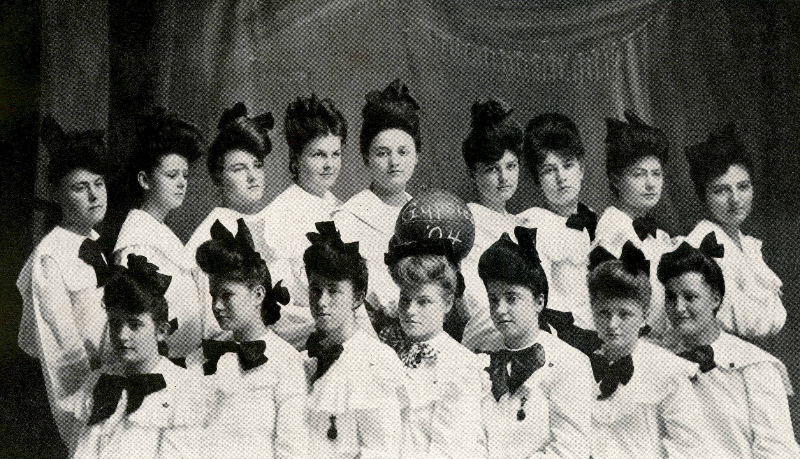 Aged team photo of the women's basketball team. They're seated in two rows, and wearing matching white blouses, dark neck ties tied in large bows, and large dark hair bows. One woman in the center of the top row holds a basketball which reads "Gypsies '04".