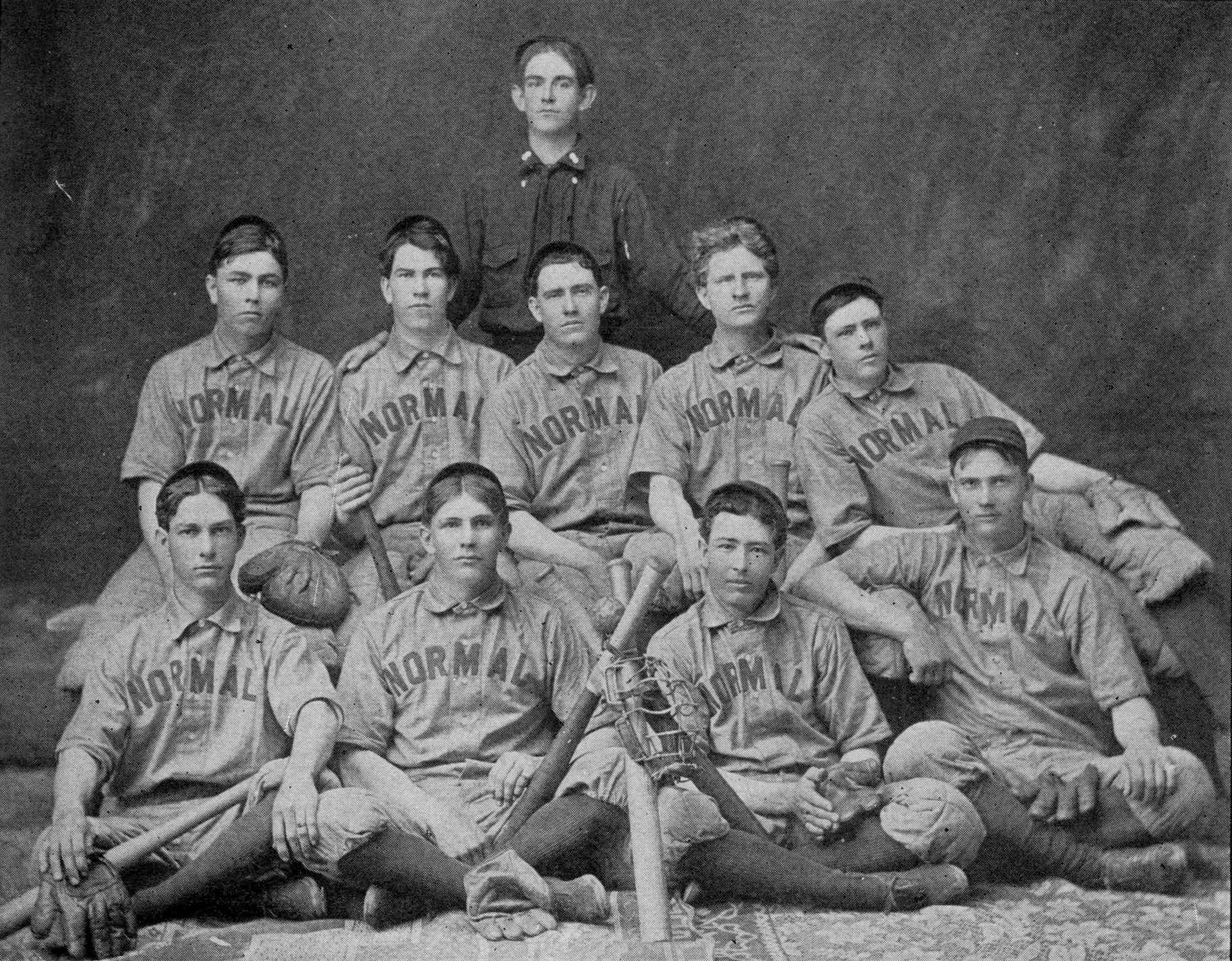 Historic black and white photo of the men's baseball team at Southwest Texas Normal School. The young men are seated in two rows, wearing their game uniforms with 'Normal' emblazoned across the chest, and dark caps. Many hold their gloves and bats. At the front of the photo, they've erected a few baseball bats into a lean-to structure and have balanced an umpire's mask on it. Behind the young men stands another gentleman with his hands on the shoulders of two of the players in front of him, wearing in a dark shirt and tie.