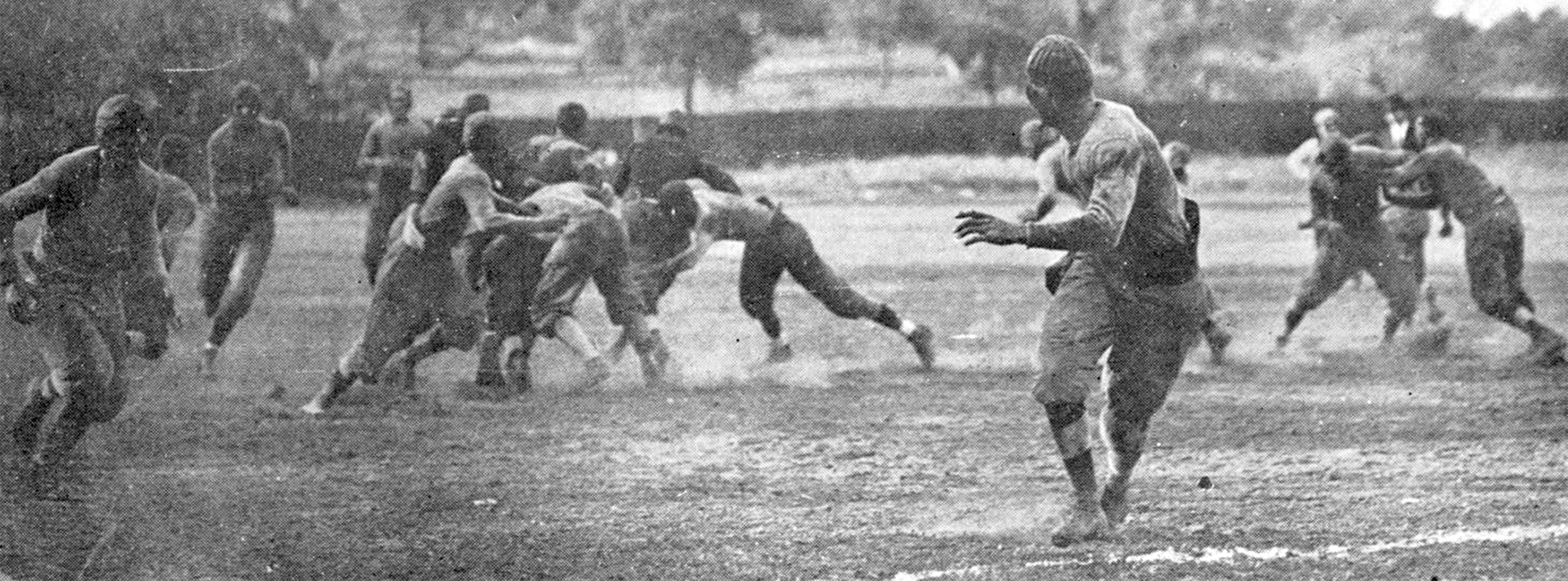 Black and white photo of several men playing a game of football in 1917. One man run towards the camera looking back over his shoulder for the ball and into the action of the game: several men are clashing in tackles, and dust kicks up around their feet. A few other men run up from behind the linemen, chasing after the foremost figure.