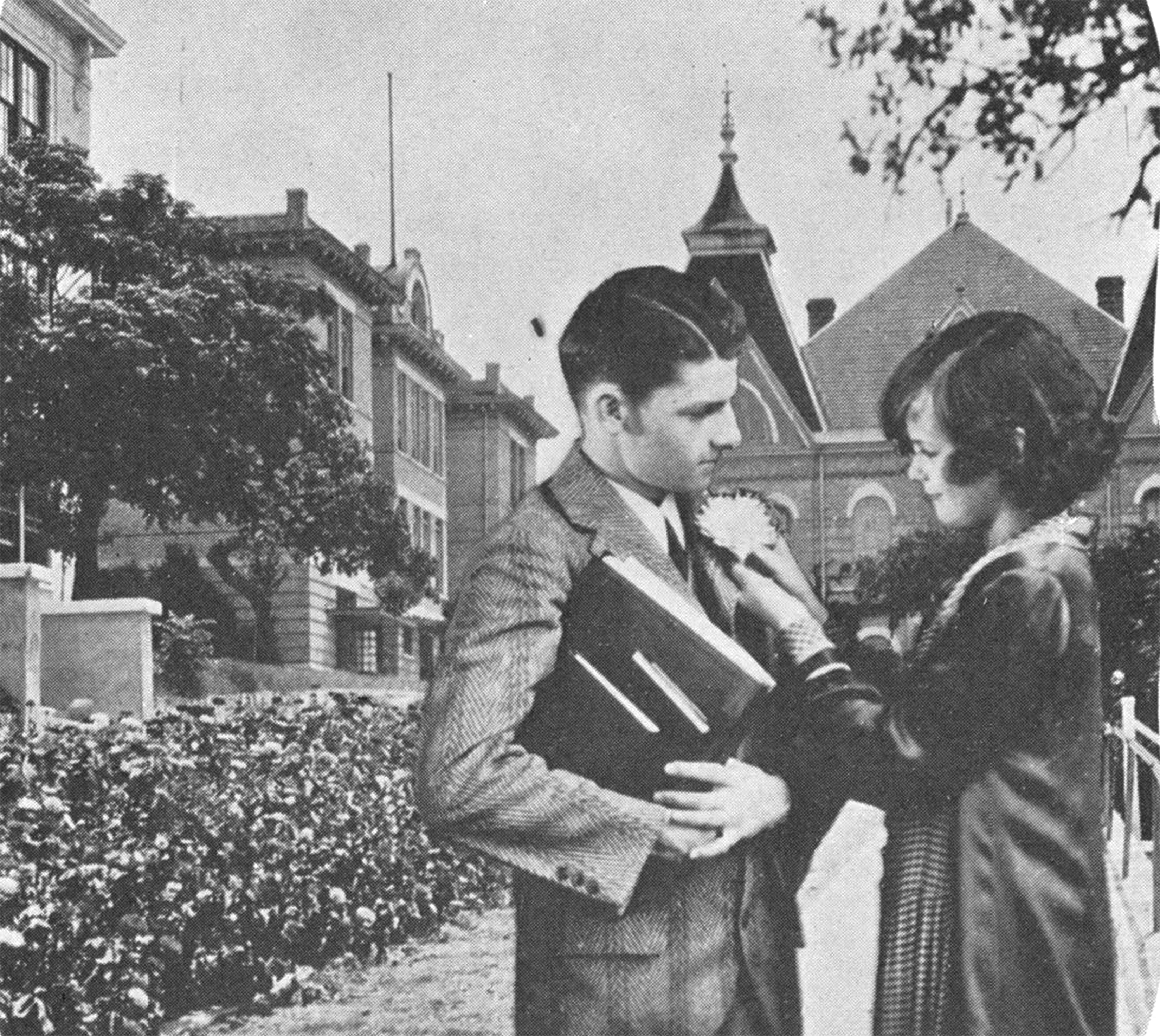 Vintage black and white photograph of a young man and woman on campus, with Old Main lording over the scene in the background. The lady is focused on pinning a large white blossom on her beau's lapel while he juggles a stack of books in his arms.