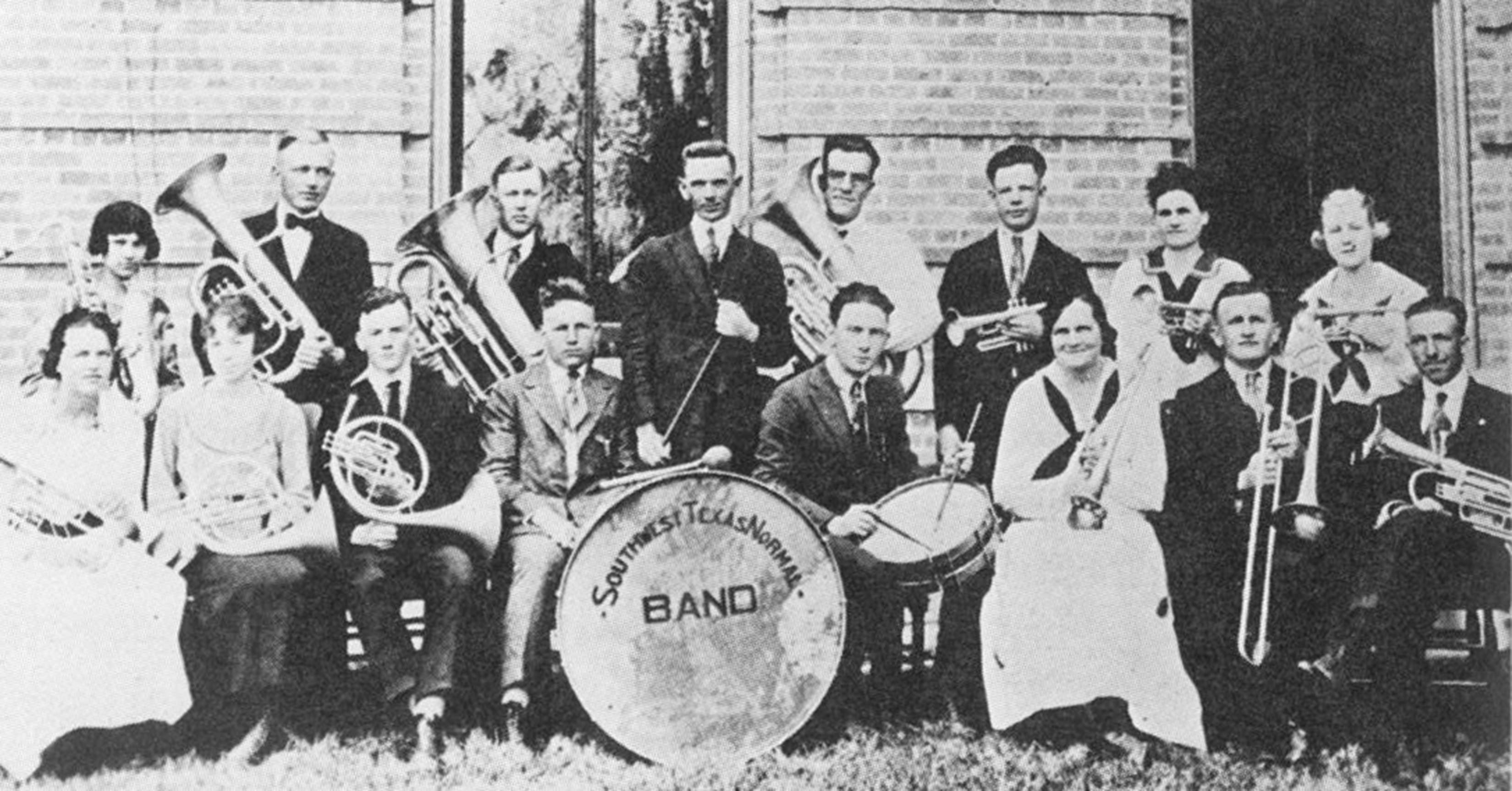 Black and white historic image of the band posing for a group photo. Arranged in two rows, the men wear dark suits and ties, and the ladies wear white dresses with dark ties done up in bows. Each young man and woman holds their instrument, including trumpets, drums, trombones, french horns, a few tubas, and even a saxophone. The band leader stands in the center holding a baton. In the center, a large drum in the middle is displayed, emblazoned with the words "Southwest Texas Normal Band."