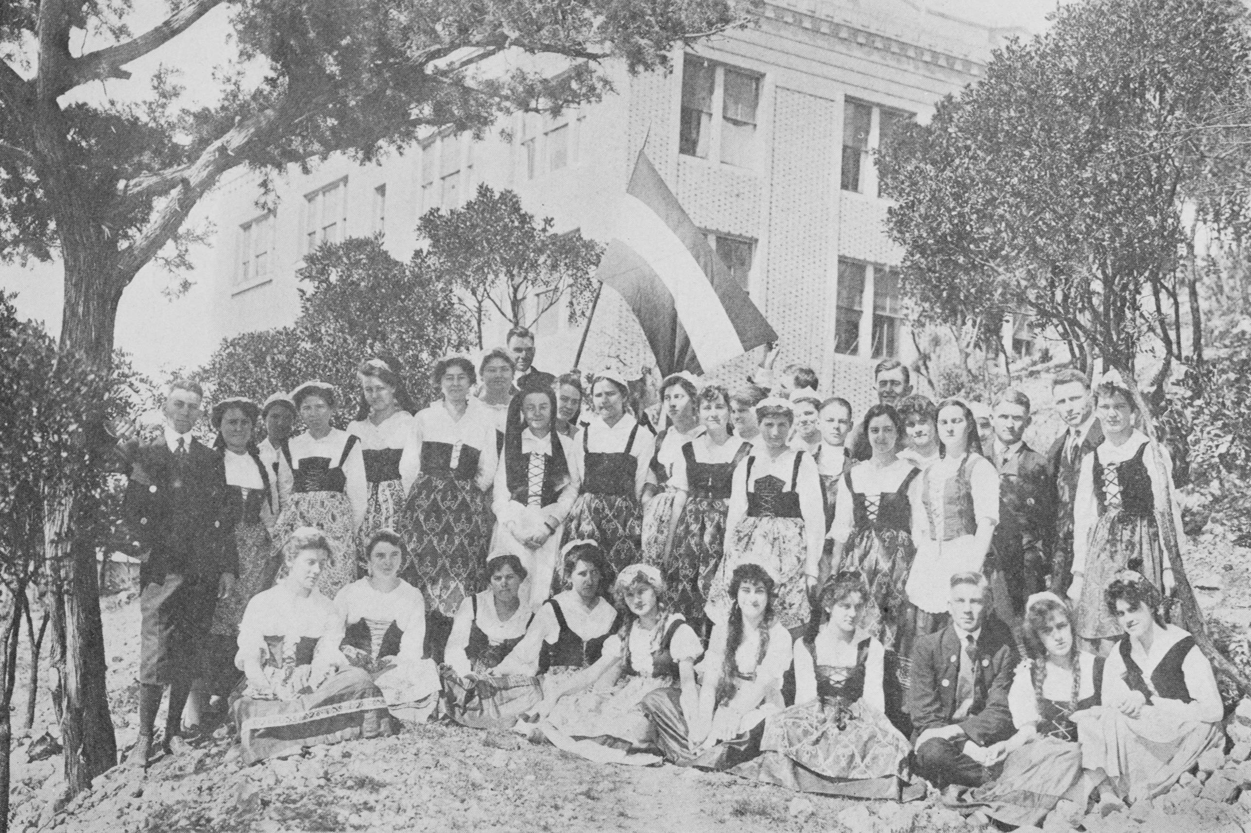 Photo of the German Club posing outside on the lawn for a group photo in 1917. The students are dressed in traditional German clothing, with the ladies in dirndls and the gentleman in jackets and short pants. A young man holds a German flag aloft in the background.