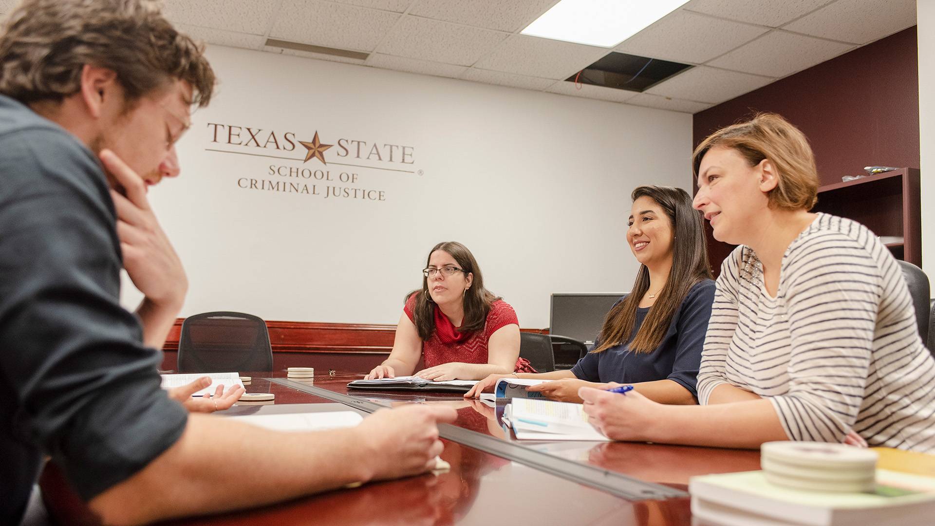 Group of four people at a conference room table with a Texas State School of Criminal Justice logo on the wall