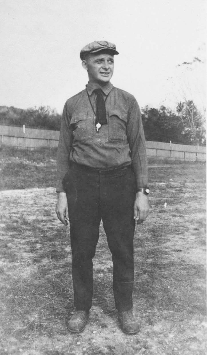 Aged black and white photograph of Oscar Strahan standing in a field and gazing right, squinting in the sunlight with his arms at his sides. He's wearing a dark button up shirt and long pants, a short dark necktie, and a flat cap with a shining whistle hung around his neck.