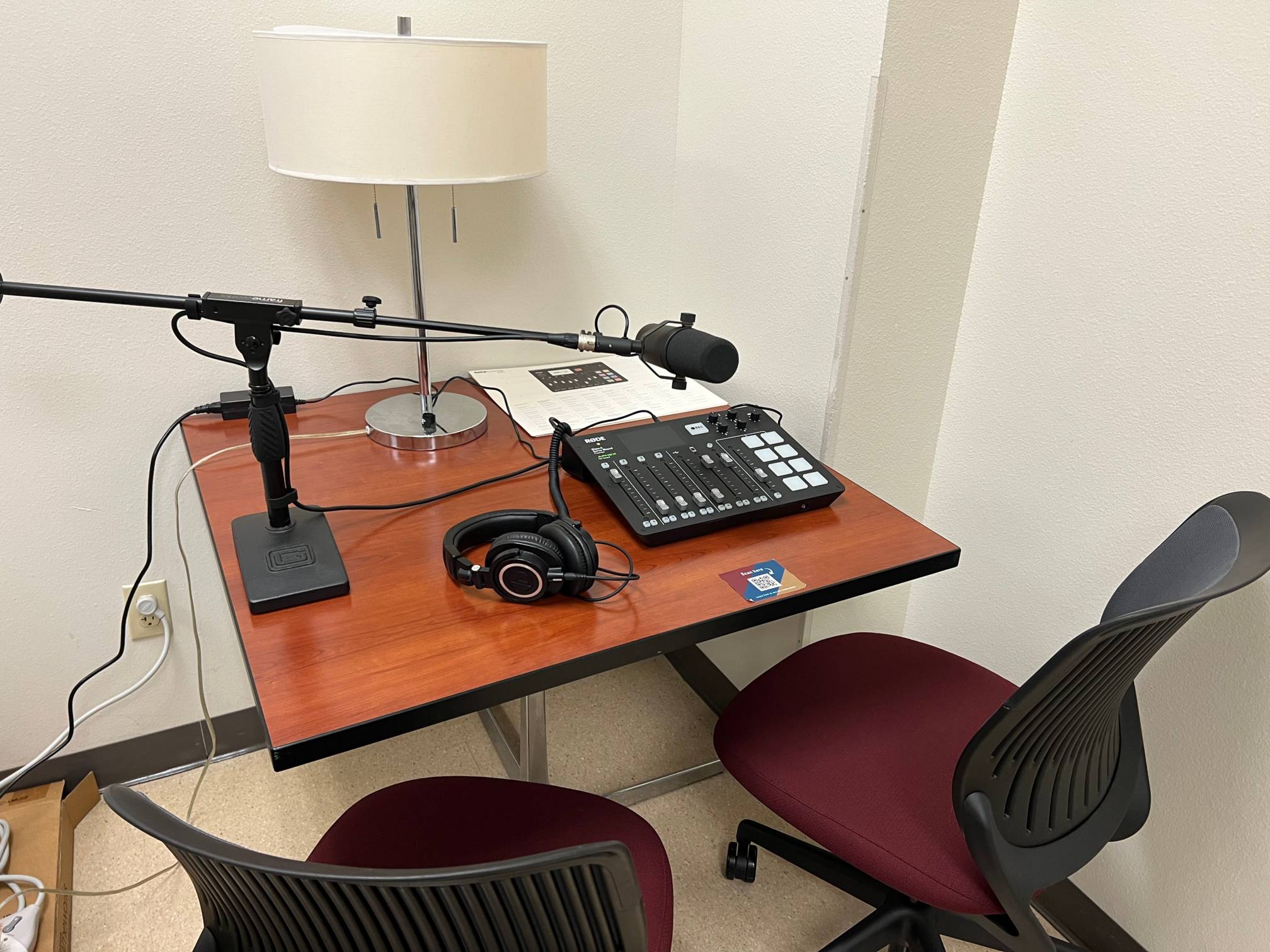 A podcast recording setup with a microphone, mixer, headphones, and lamp on a wooden table, accompanied by two chairs