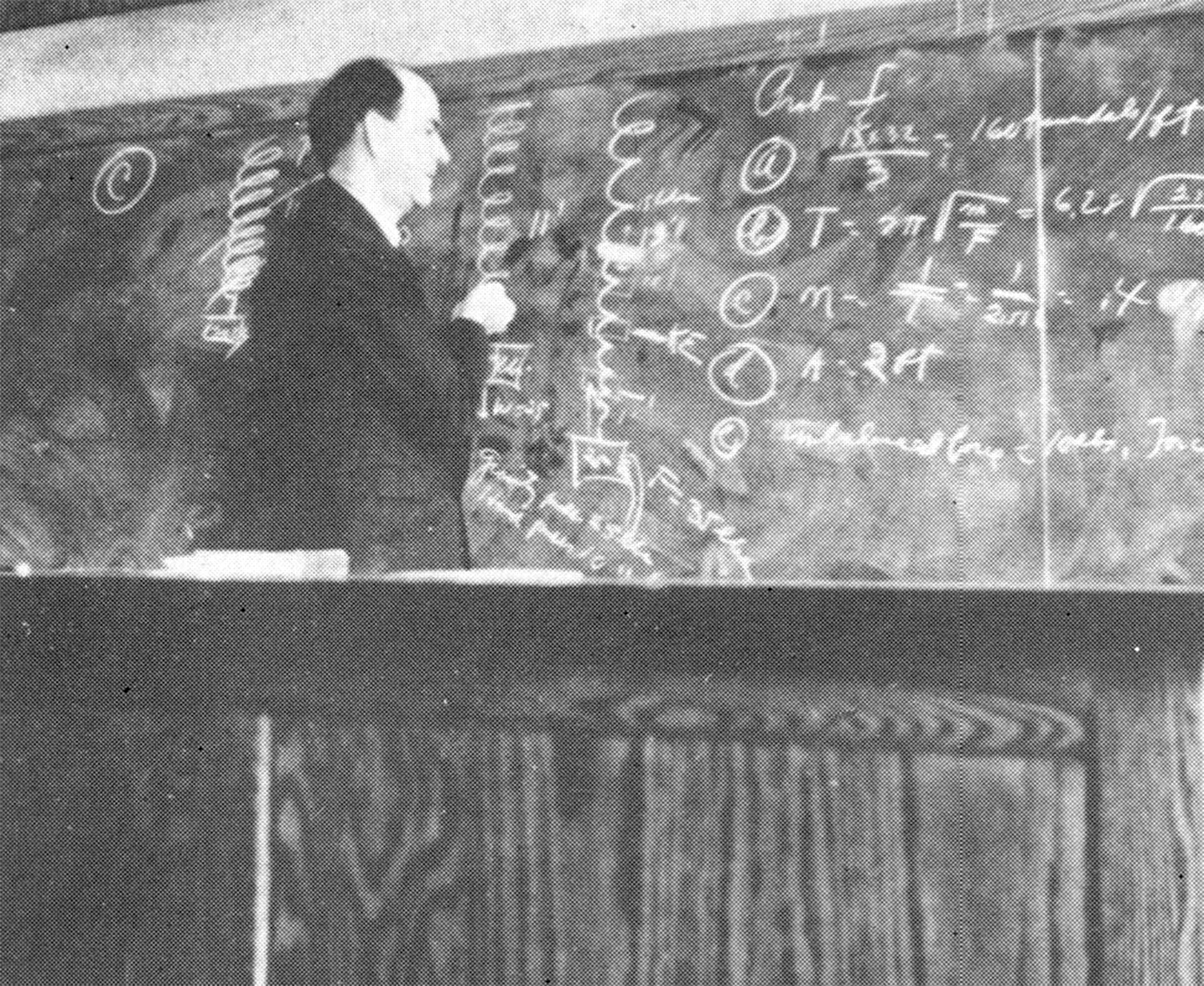 Black and white snapshot of a university professor in 1938. A balding man in his middling years, he stands behind a heavy wooden desk and is writing on a large chalkboard covered in mathematic formulas and diagrams.