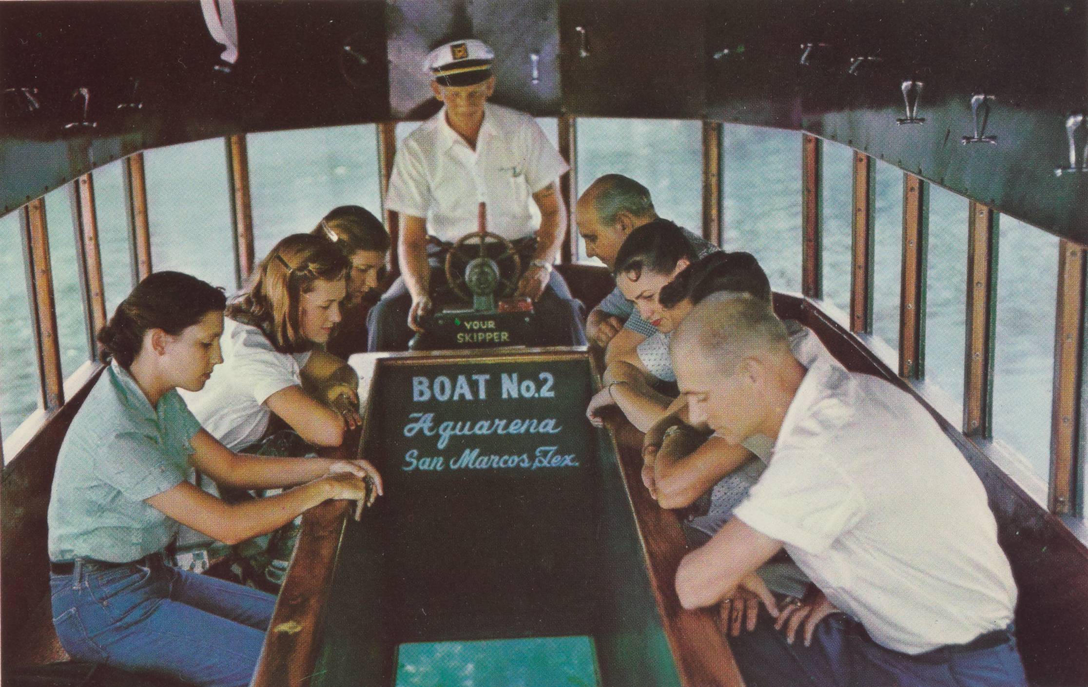 An interior snapshot of one of Spring Lake's early glass-bottom boats. A cluster of people are seated on each side of the walled glass window at the bottom of the boat, leaning on the railing to look down into the clear blue water. At the head of the group, the boat's skipper is seated and narrates the journey for his passengers. On one wall is painted 'Boat No. 2. Aquarena. San Marcos, Tex.' 