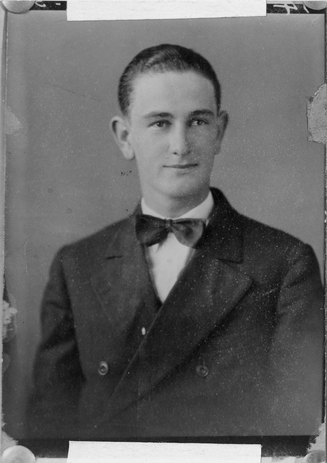 Aged portrait of a young Lyndon B. Johnson when he was a college student. Wearing a dark, neat suit with a bowtie, his hair is slicked back and he smiles slightly for the camera. 