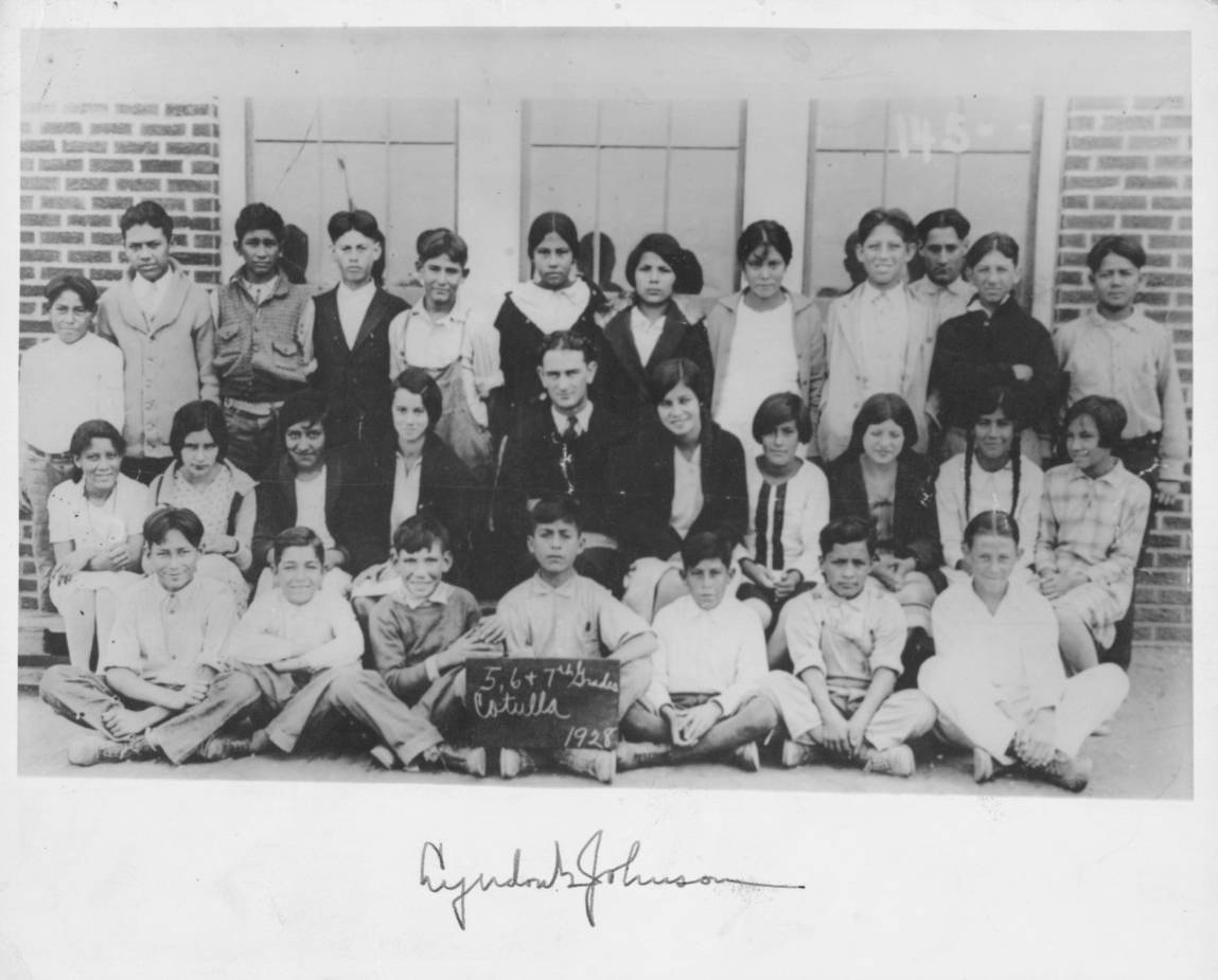 Black and white class photo of Lyndon B. Johnson with his pupils. The class consists of twenty-eight students, and the students are lined up in 3 rows, the back stands, the middle in chairs, and the bottom row seated cross-legged on the floor. As the teacher, Johnson is seated in the center of the middle row, arms folded. The student in the center of the bottom row holds up a small chalkboard sign reading '5, 6, and 7th Grades. Cotulla. 1928.'