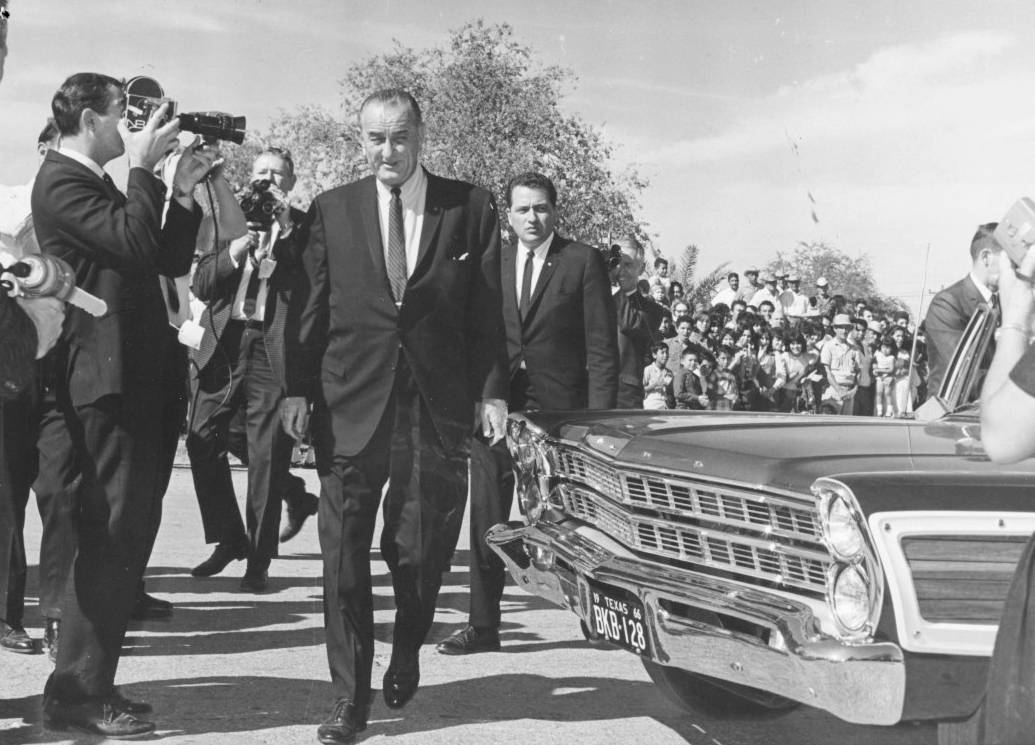 Aged snapshot of Lyndon B. Johnson as president. President Johnson smiles as he crosses in front of a vintage car with Texas plates, closely followed by a Secret Service agent. Several photographers stand to his left and around the president, snapping photos as he passes. In the background, a large crowd of men and women observe the scene with interest.