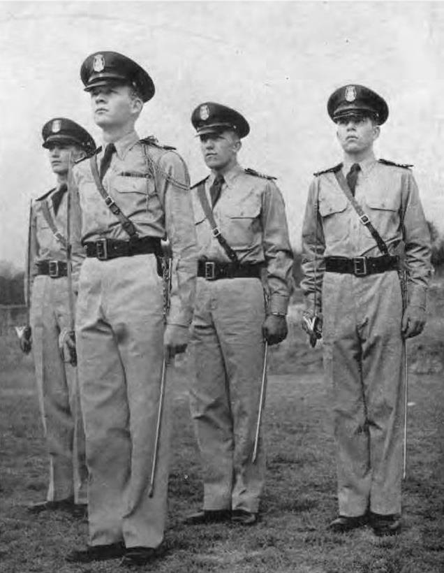 Vintage portrait of four Texas State students, standing at attention and wearing their dress uniforms for the Air Force ROTC program. Each wears a short light jacket with a matching color shirt, pants, and a dark neck tie. They wear large, dark belts, with a leather strap crossing from their right shoulder to left hip, and each carries a sheathed military saber. Each also wears a peaked cap with the company's insignia, and shoulder marks. The man in the front also has a braided cord signifying rank hanging from his right shoulder.