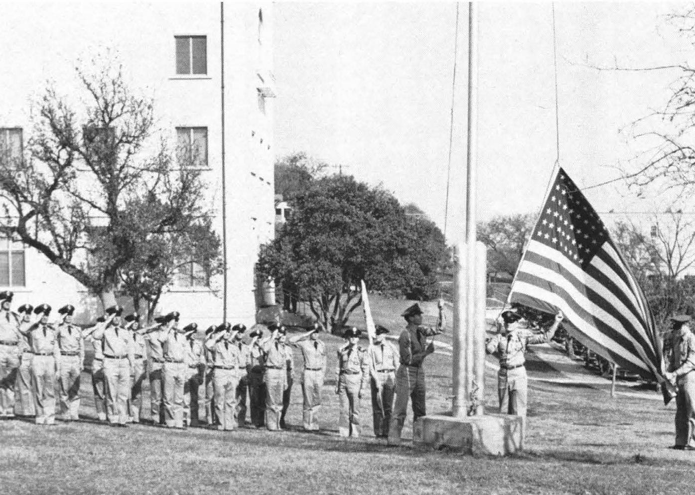 Aged black and white photo of Texas State Air Force ROTC cadets raising a United States flag on campus. One young man handles the pulleys to raise the flag up the pole, while two more ensure the flag does not touch the ground. Behind them, the remaining cadets stand at attention and give a salute as is custom.