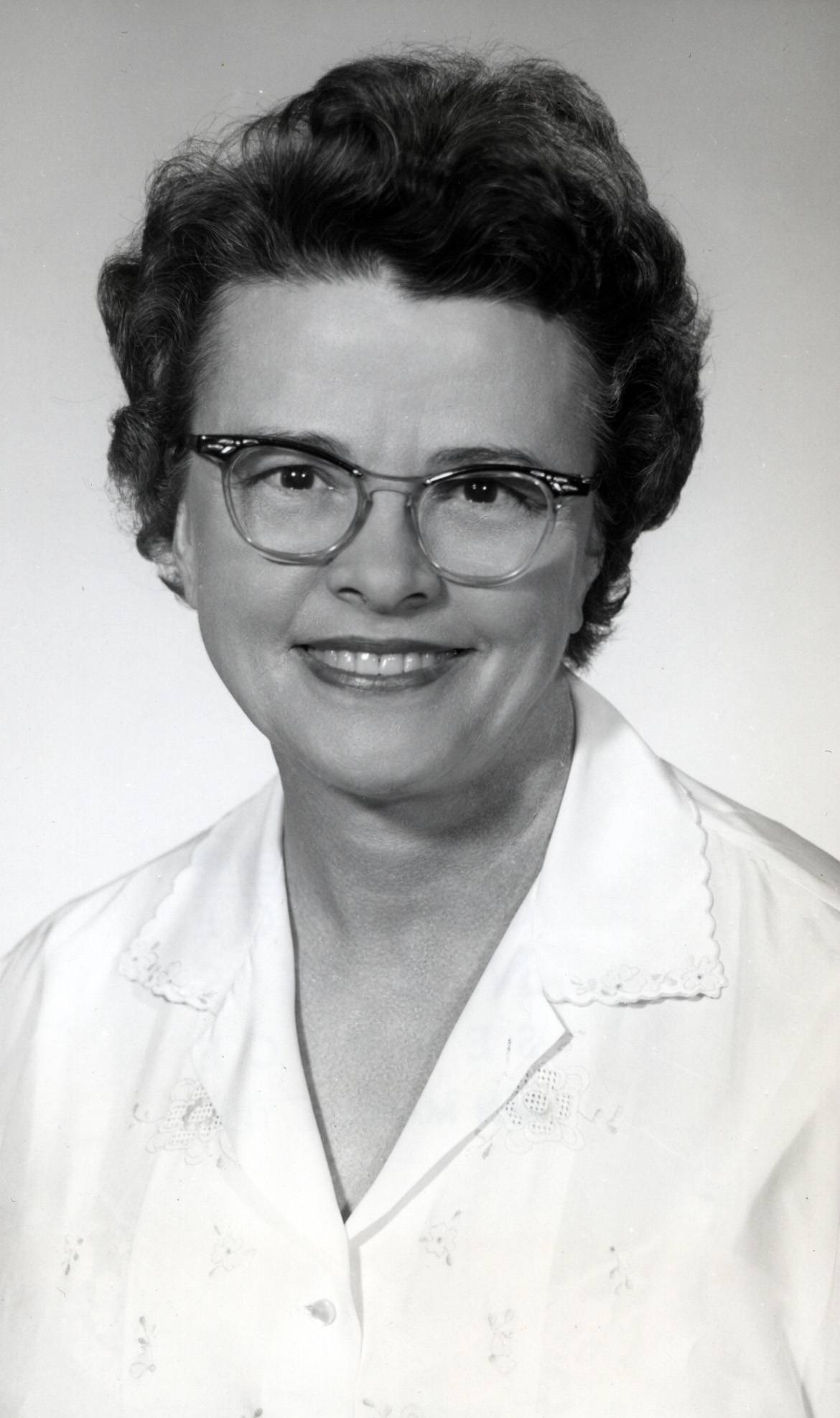 Black and white headshot of Dr. Empress Zedler in the late 1940's or early 1950s. She's fair-skinned woman with dark eyes, and dark, wavy hair cut short. She appears to be in her middle years, wearing stylish cat-eyed glasses, and a white collared shirt decorated with simple, floral embroidery.