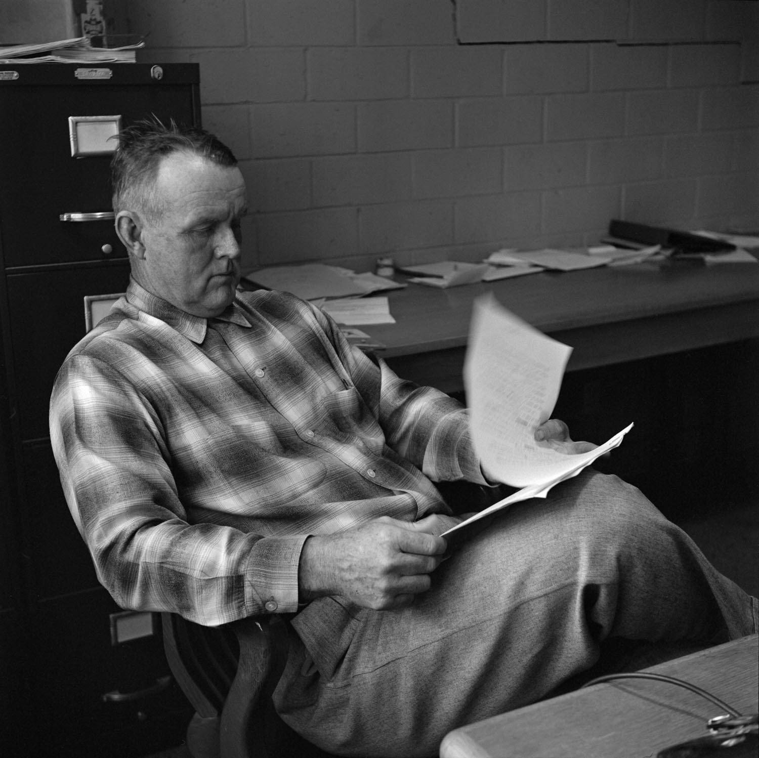 Snapshot of Milton Jowers during his time as a coach at Texas State. In his middling years, he has short graying hair cropped short, and wears slacks and a flannel shirt. He reclines in his seat with his legs crossed as he flips through a stack of papers, concentrating on his task.