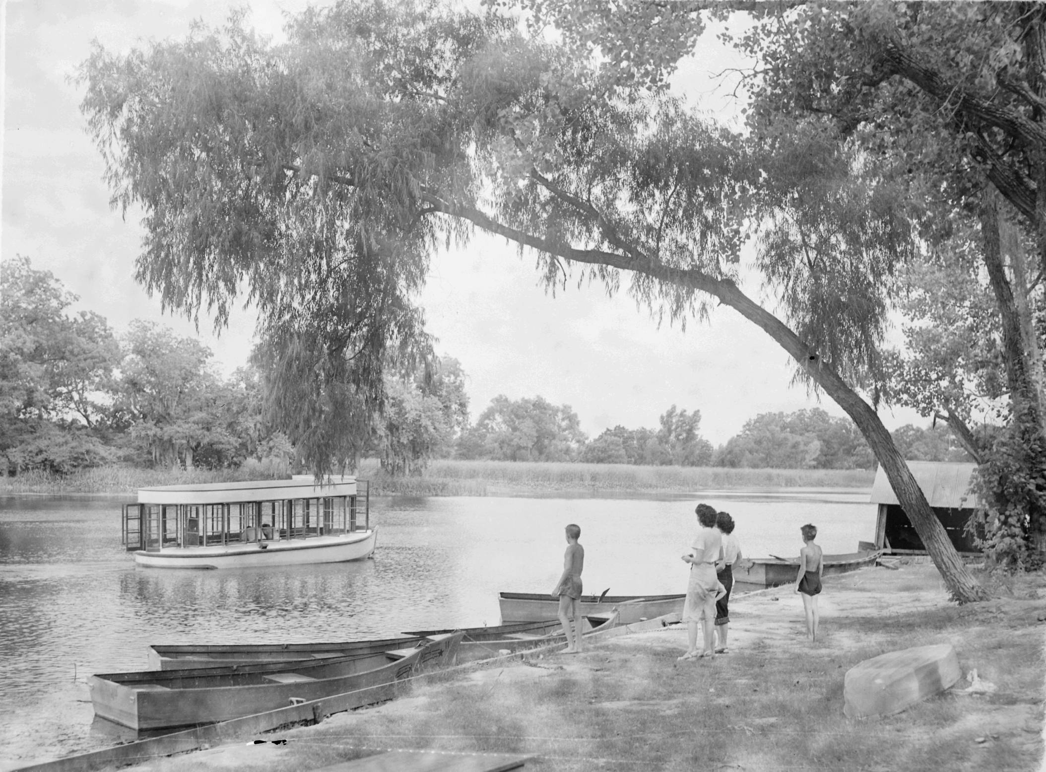 Black and white photograph showing a vista around Spring Lake. One of the famed glass-bottom boats sails past serenely. Small paddle boats are docked along the shore, and a cluster of four swimmers stand in the shade of tall trees on the shore, enjoying the view.