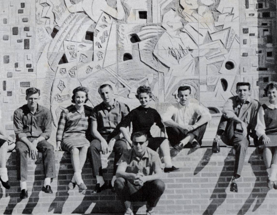 Black and white photo of students posing for a group photo in front of the iconic Buck Winn mural. The mural is a relief decorated with pieces of glass on each side and dotted throughout the composition. The figures in the piece are slightly abstracted, simplified and geometric, reminiscent of art deco and cubism.