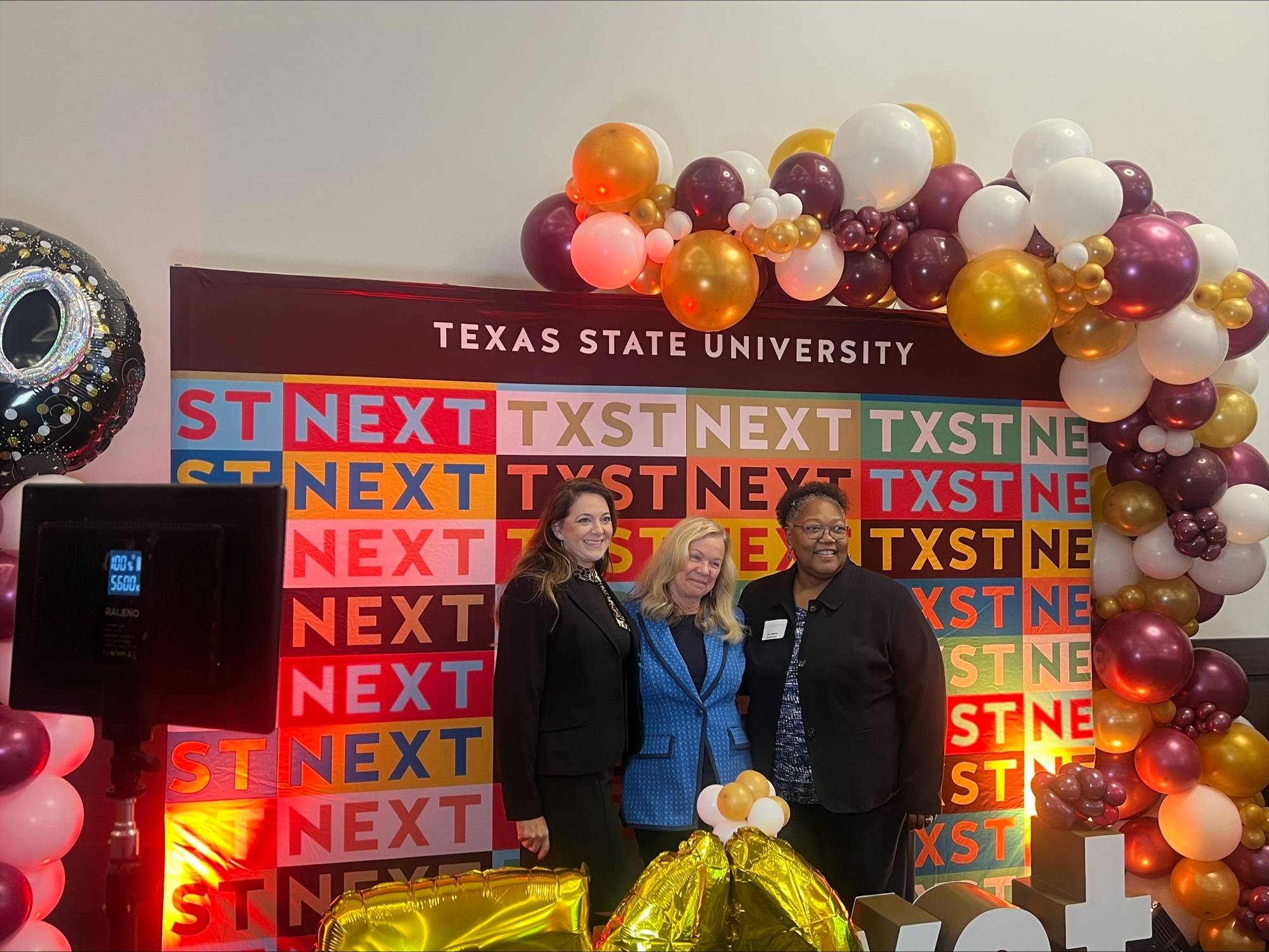 Three people in business clothing pose in front of a colorful backdrop, surrounded by balloons.
