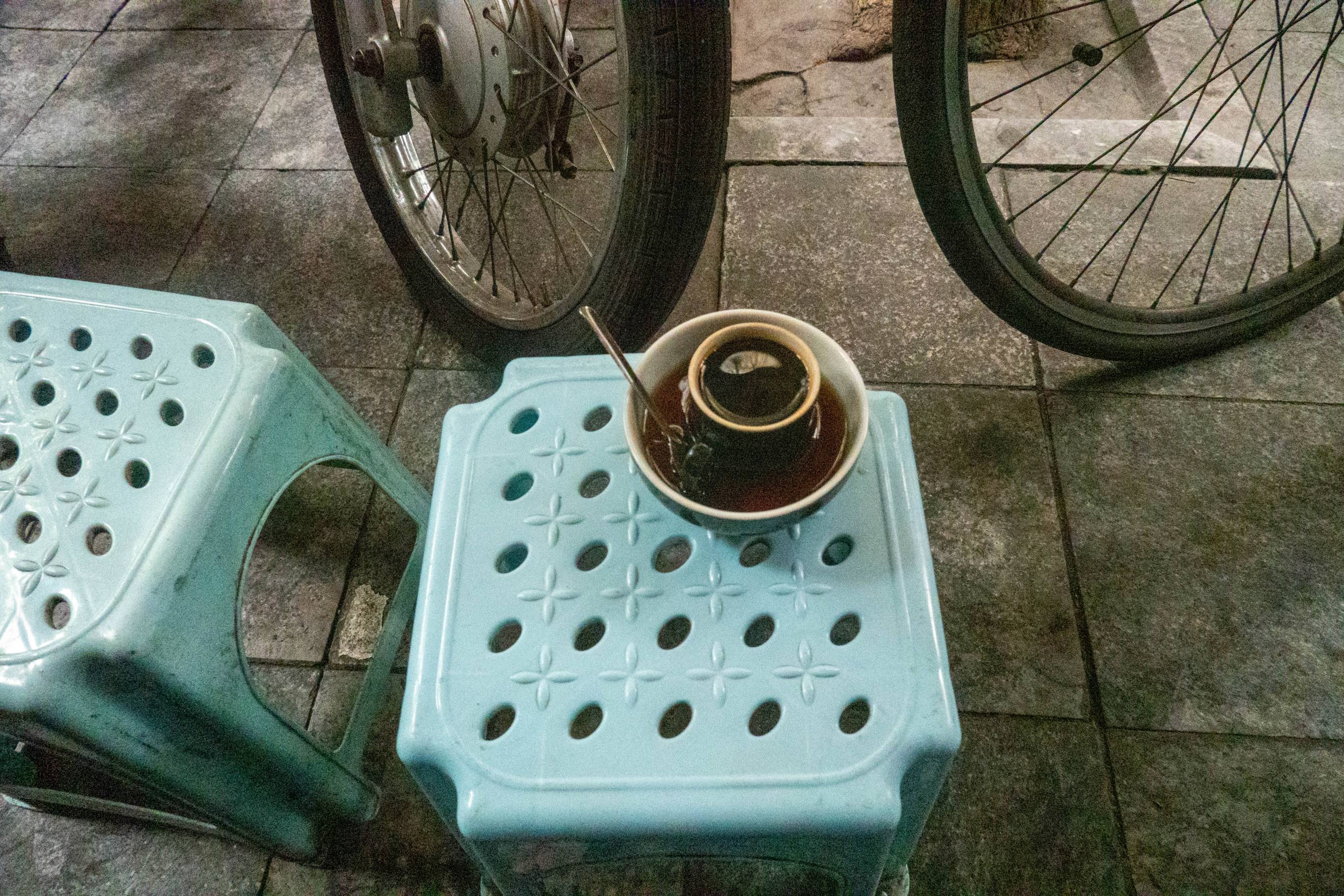  hot black coffee in a porcelain cup and then put that cup into a bowl of coffee and ice sitting on a small blue plastic table in front of two bike tires