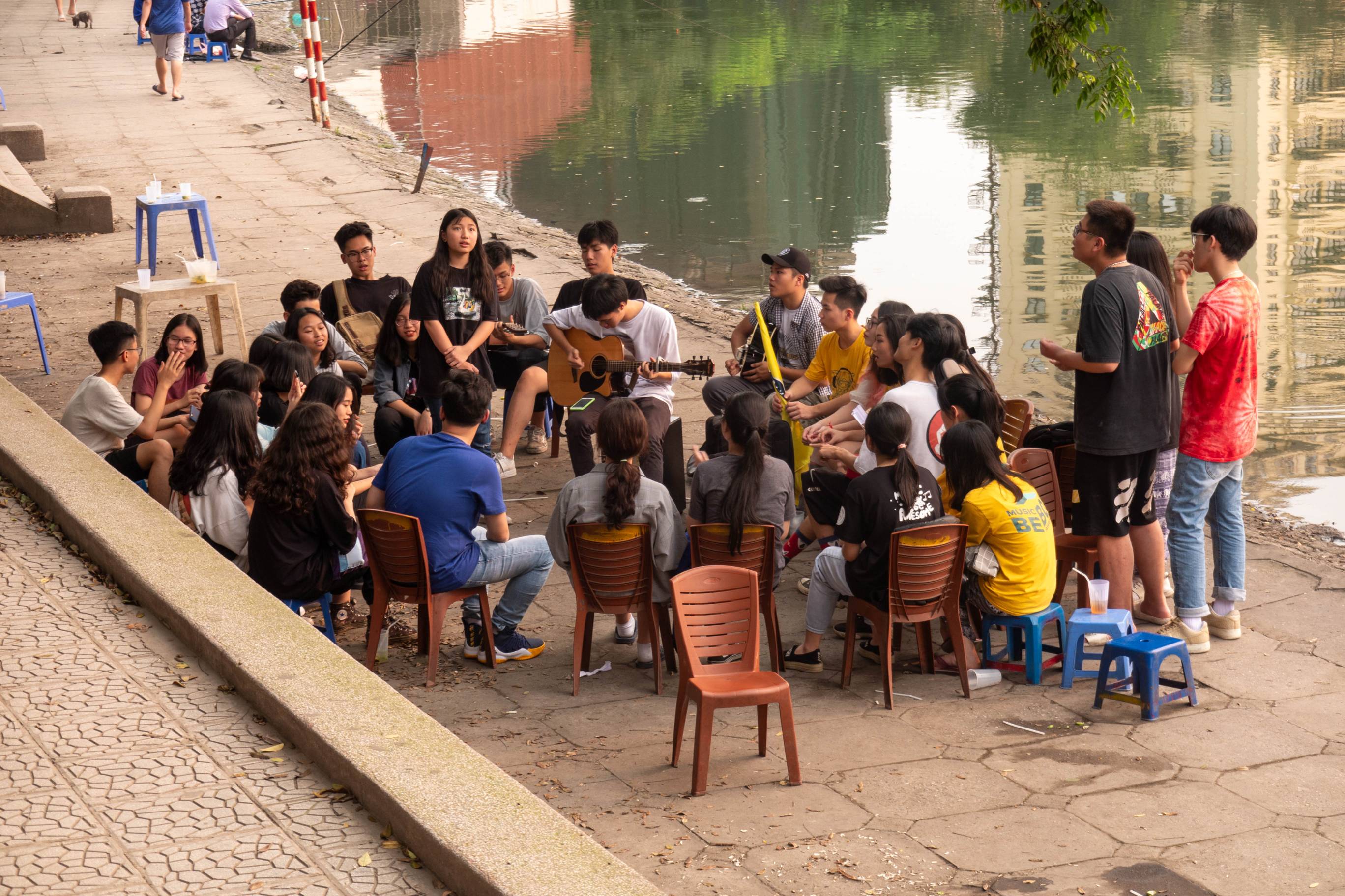 Picture of young man at a lake in the center of a group of young people playing an acoustic guitar.