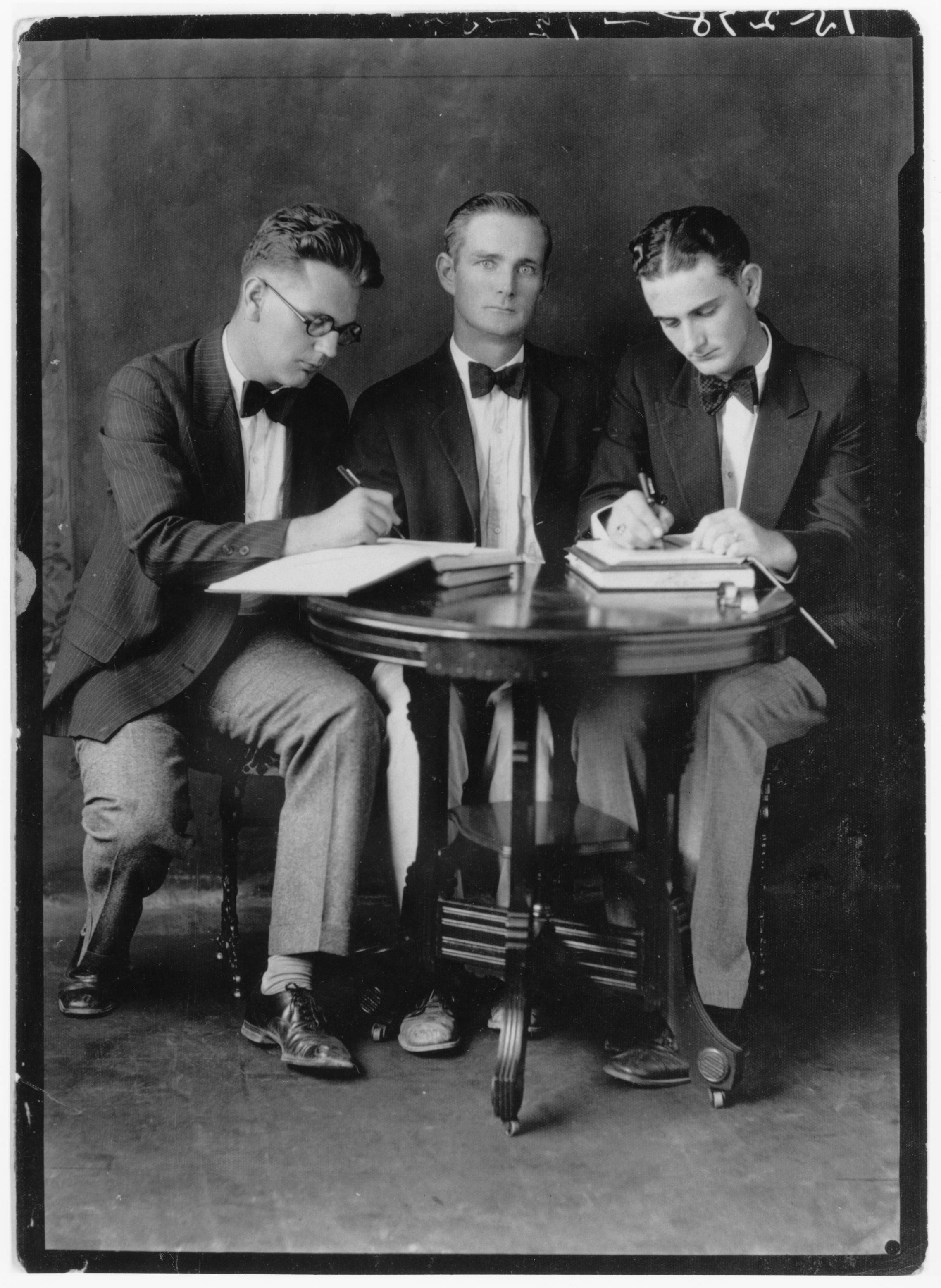  A black and white photo of Lyndon B. Johnson sitting alongside two other young men. All three are wearing suits with beige pants, dark jackets, and bowties. The gentlemen in the middle stares intensely at the camera while LBJ and the other Bobcat appear to be focused writing into paper tablets. LBJ is looking downward as he writes with a calm expression.