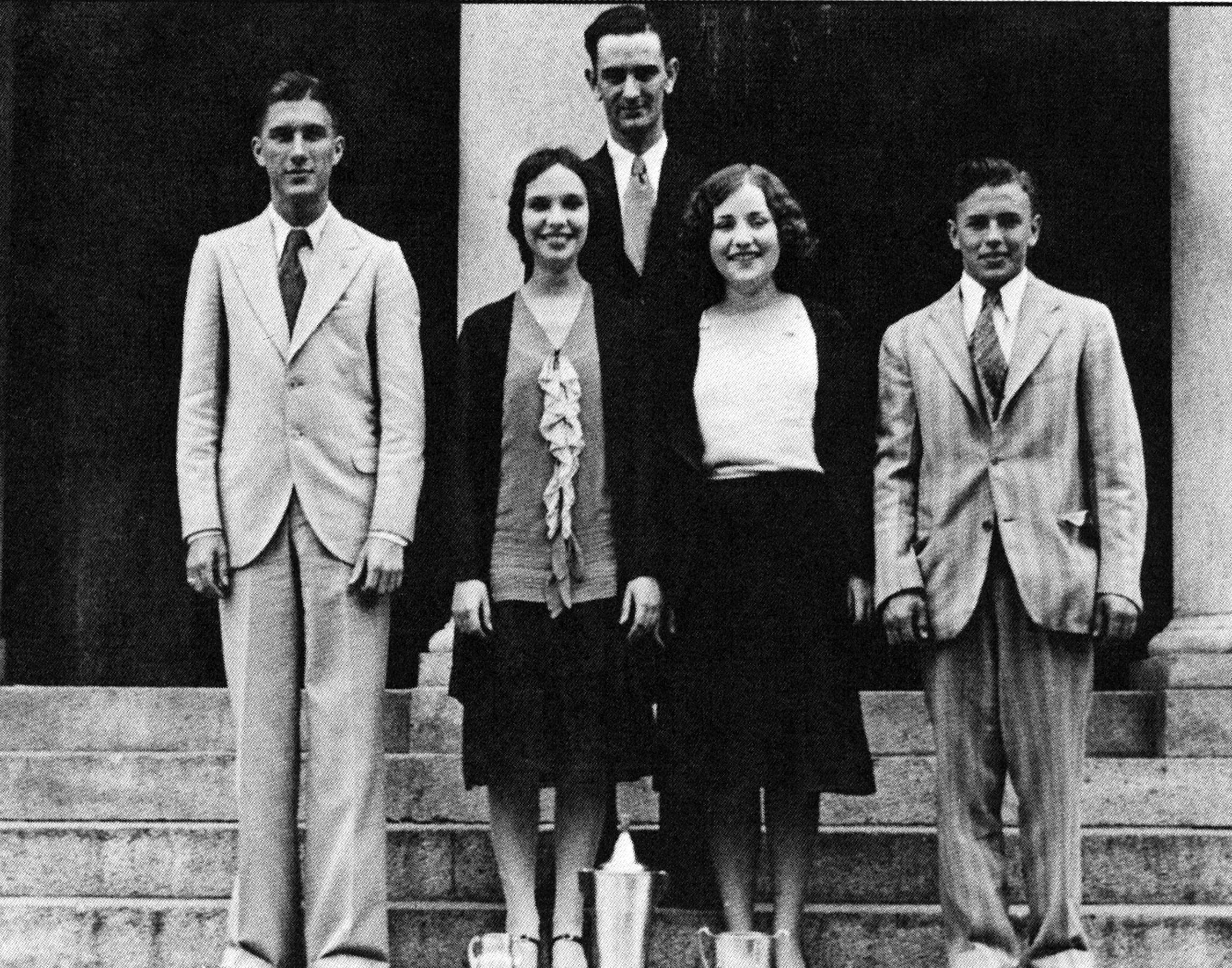 A college-aged Lyndon B. Johnson, dressed sharply in a dark suit, stands alongside four other Texas State students whom are all dressed immaculately in suits and professional wear. The young adults are smiling slightly as they pose for the photo. 