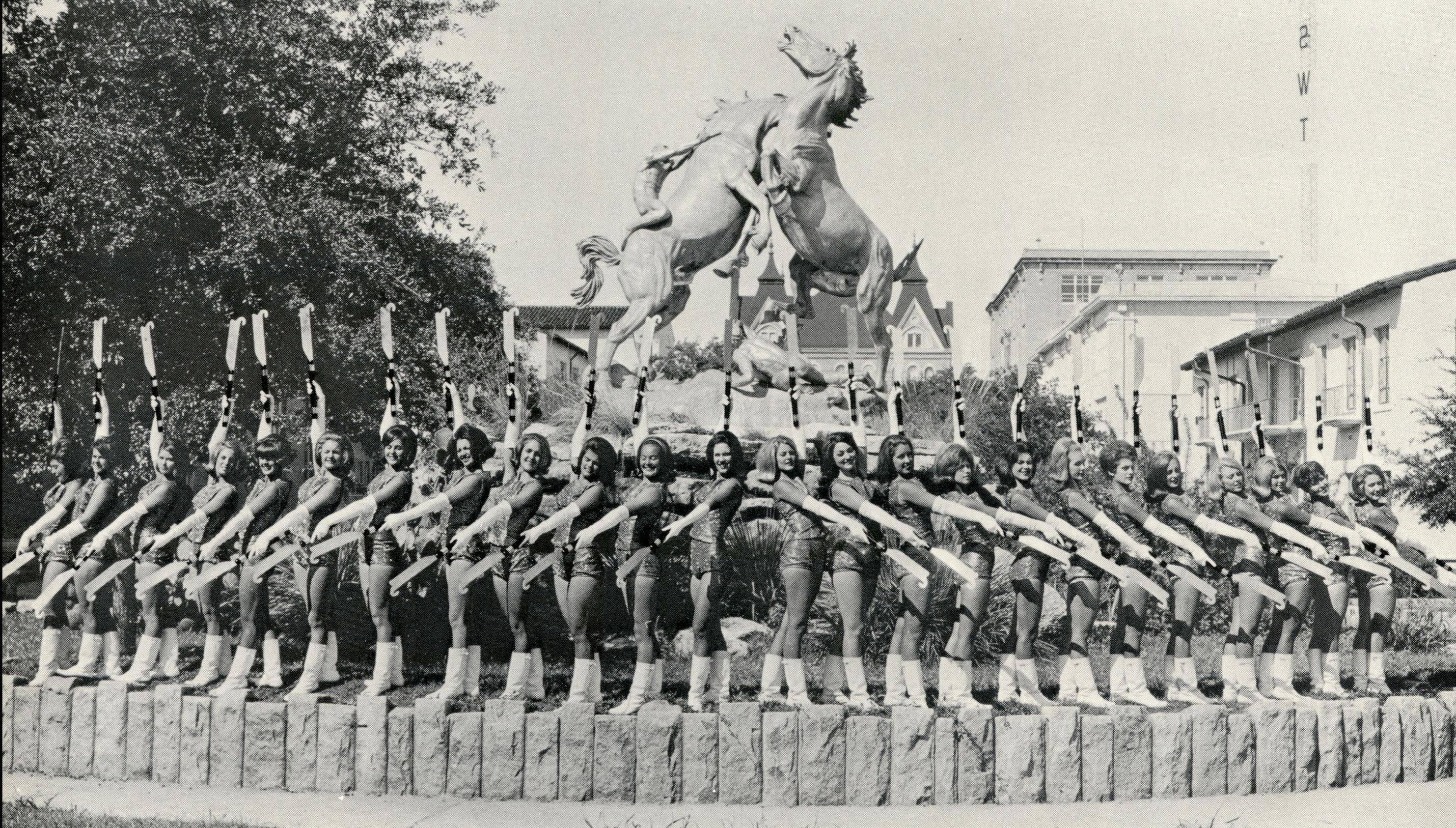 A large group of Strutters are perched on a landing just below the iconic Fighting Stallions statue. The group looks onward in matching uniforms as they hold up their batons. In the background can be seen Old Main and other historic San Marcos Campus buildings. 