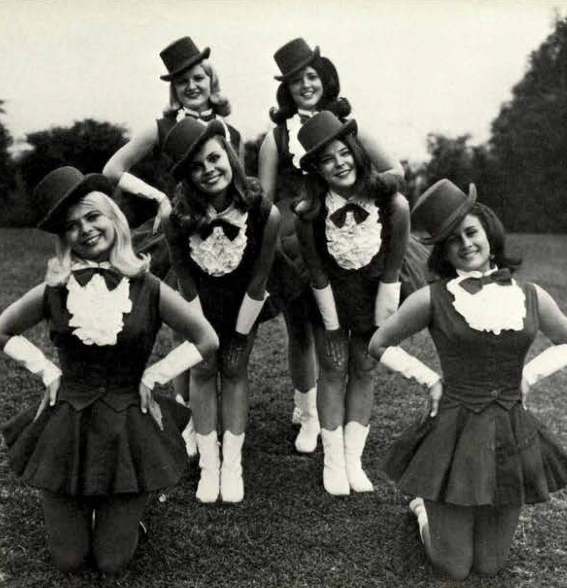 Six Strutters smile brightly as they pose outdoors. The six are wearing matching attire that include ruffled tops, bowties, and top hats. 