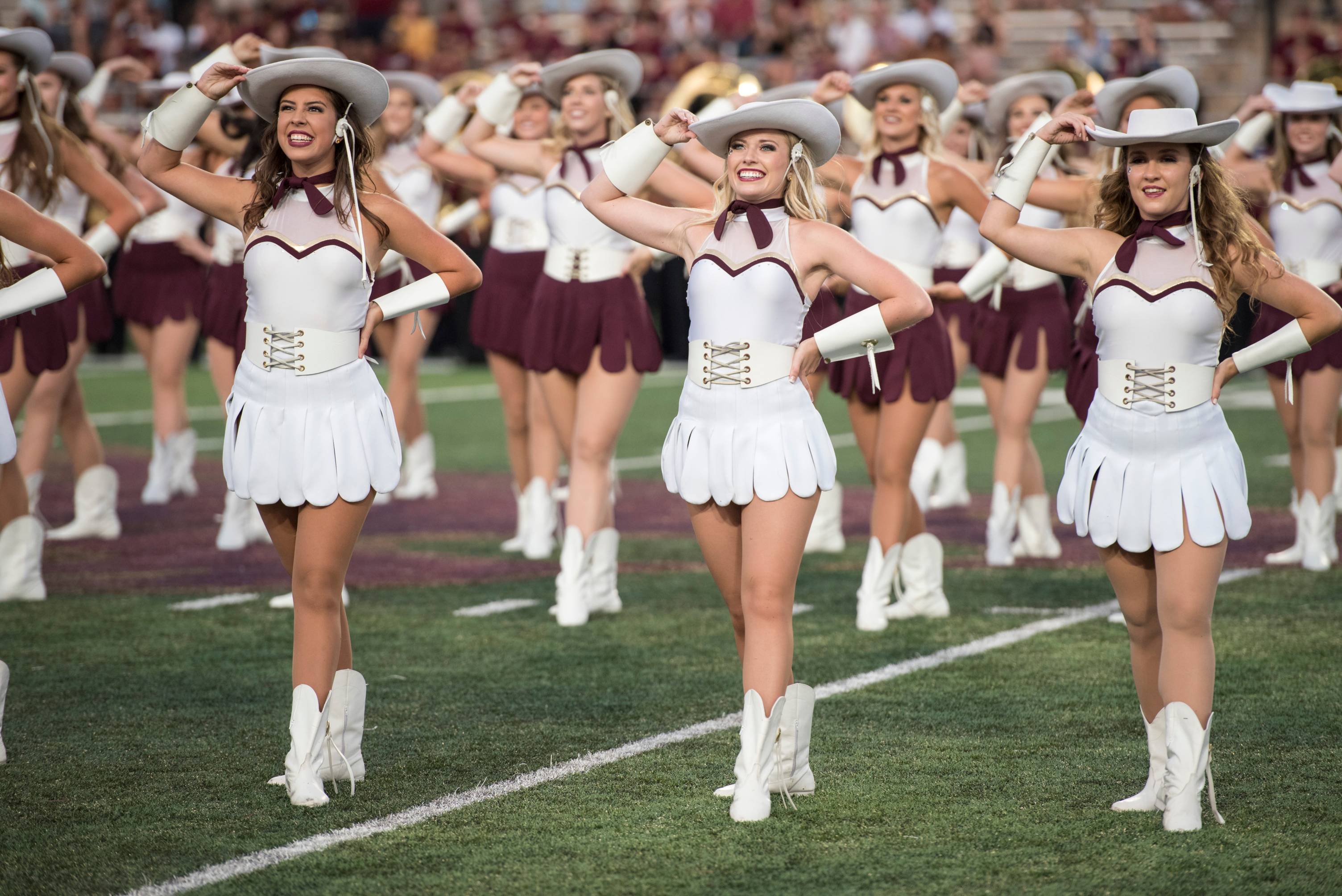 A modern photo of Strutters beaming upwards as they perform for a packed crowd at Bobcat Stadium. The dancers are all holding their cowboy hats with their right hands in anticipation of their next move in their routine.  