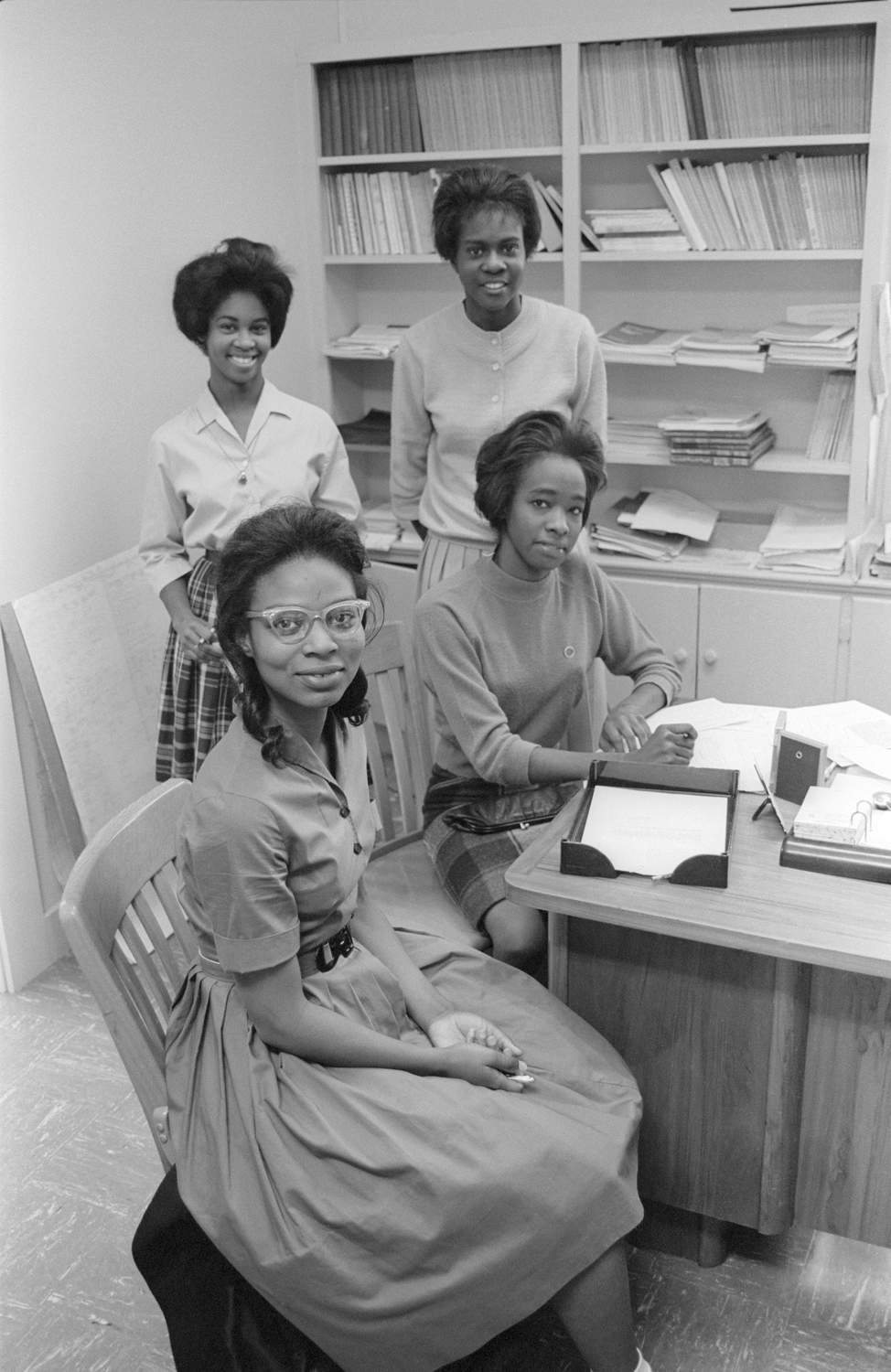 Four of the first five Black students to attend TXST register for classes. The young women are smiling in anticipation of breaking down a meaningful barrier. Shelves in the background contain books and documents.