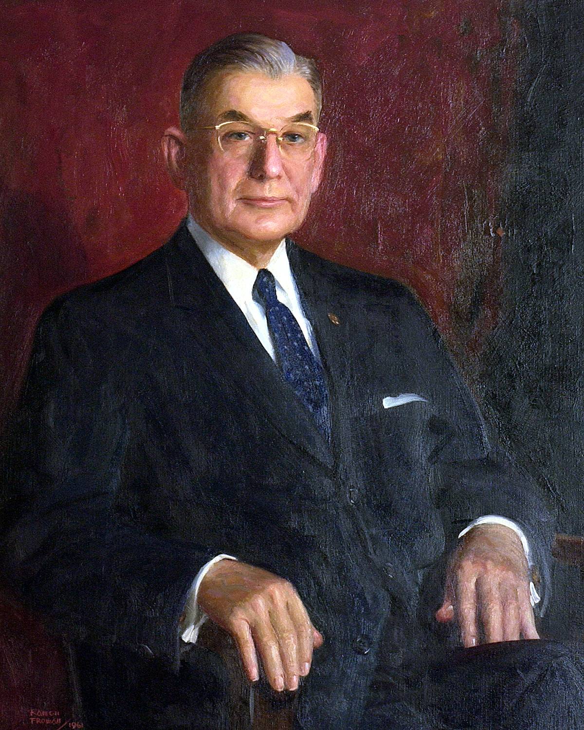 A painted portrait of the third Texas State president, John Garland Flowers. The first native Texan and first TXST alumnus to hold the presidency, Flowers is portrayed as a middle-aged man in a dark suit wearing golden spectacles. He is clean shaven and has a slight smile on his lips.