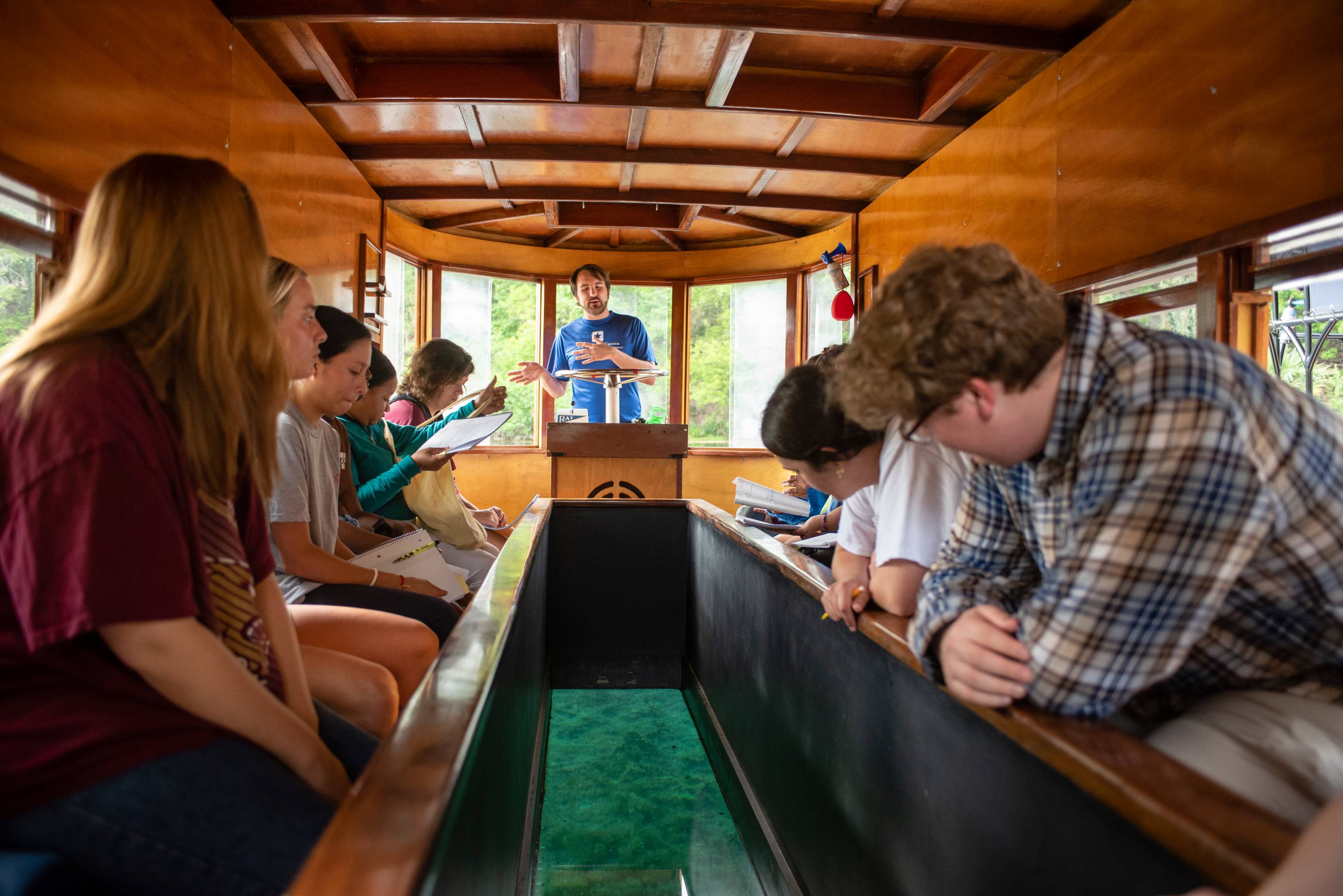 A modern photo of TXST students observing Spring Lake as their boat driver highlights aspects of the beautiful scenery below.  