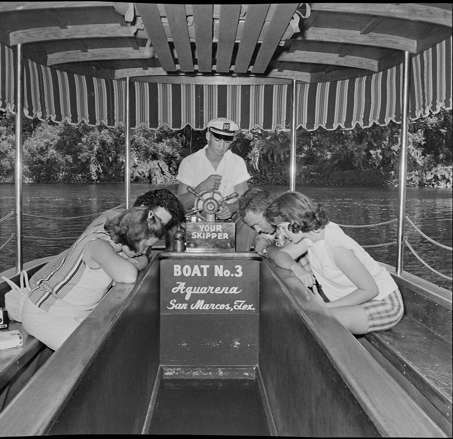  A black-and-white photo of visitors to Aquarena Springs riding in a glass-bottom boat. The young driver wears a captain hat while the riders look downward into the spring via a glass panel.  