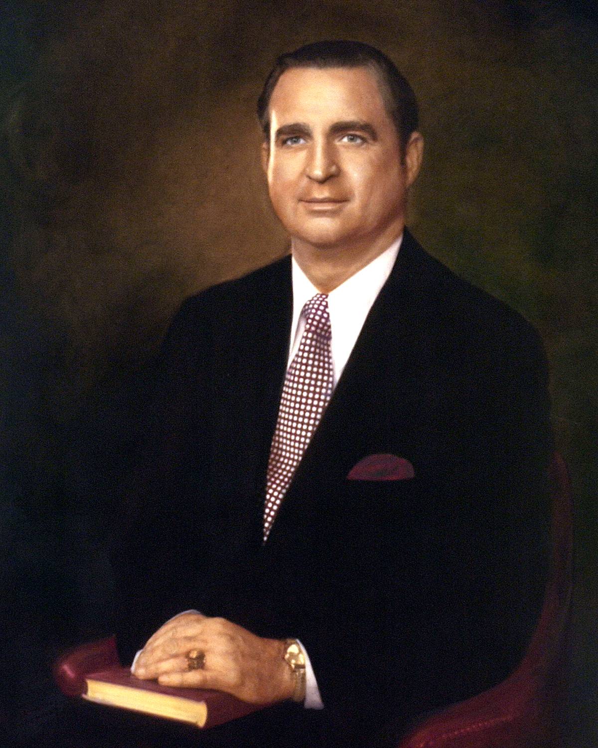 Painting of Billy Mac Jones. He's a fair-skinned man with neatly combed, light brown hair and blue eyes. Seated in a simple, reddish-leather chair, he's wearing a dark suit with a checked red tie and deep red pocket square. His hands are folded on top of a slim, leather-bound book. He's also wearing a gold signet ring, and a gold watch peeks out from under the cuff of his jacket.