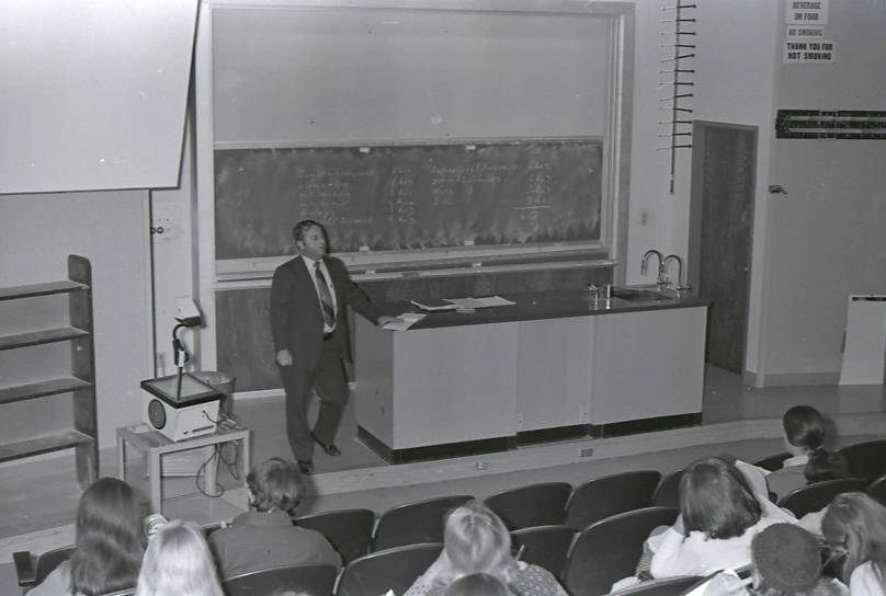 A professor appears deep in thought standing at a table with a chalkboard behind him as he delivers a lecture. An overhead projector can be seen in the image along with the tops and back of heads.