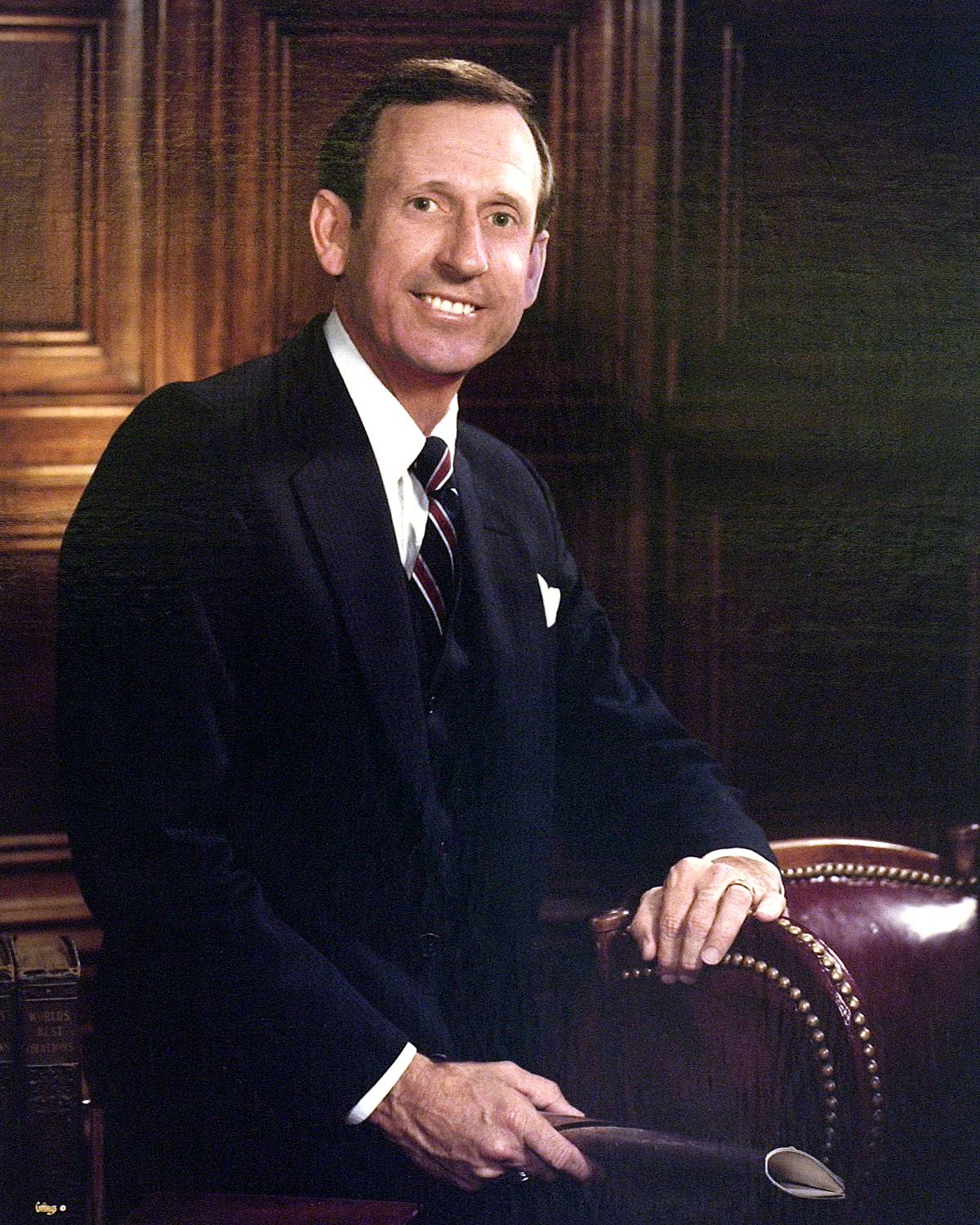 A very jovial portrait of TXST President Lee H. Smith. He has a large smile on his face while his left hand is situated on a gorgeous leather chair beside him. He’s wearing a handsome suit with a beautiful striped tie as he stands in front of a wooden paneled wall. 