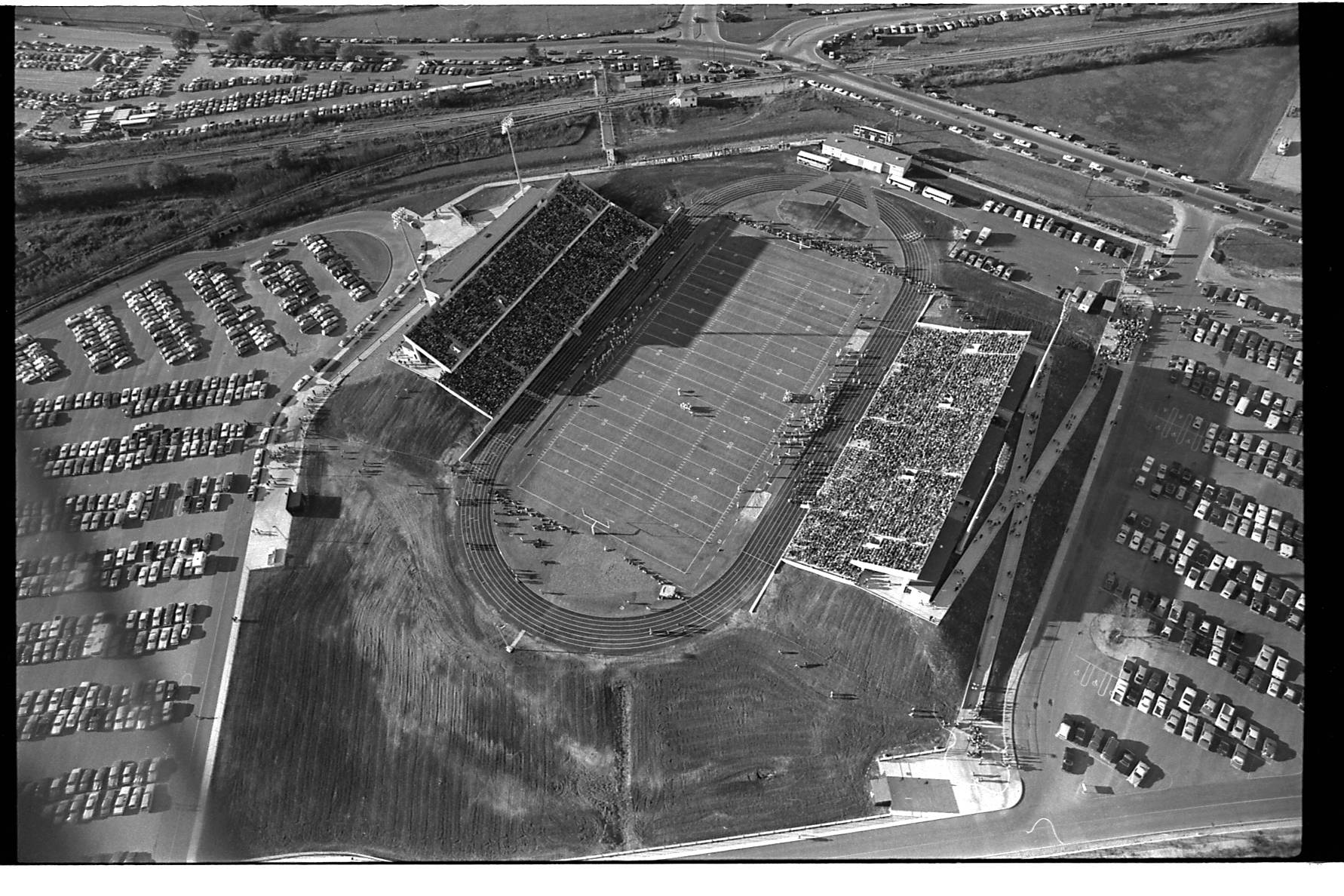 A black and white aerial photo looking down over a football stadium with parking lots full of cars surrounding it. The seats of the stadium are all filled and a football game is being played on the field.