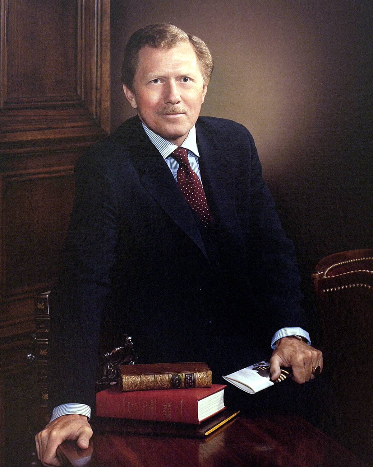 A portrait of former Texas State President Robert L. Hardesty, depicted wearing a dark suit, a blue dress shirt, and a maroon tie with polka dots. He is seated beside a stack of several books, with his right hand resting neatly next to them, while his left hand holds an open book.