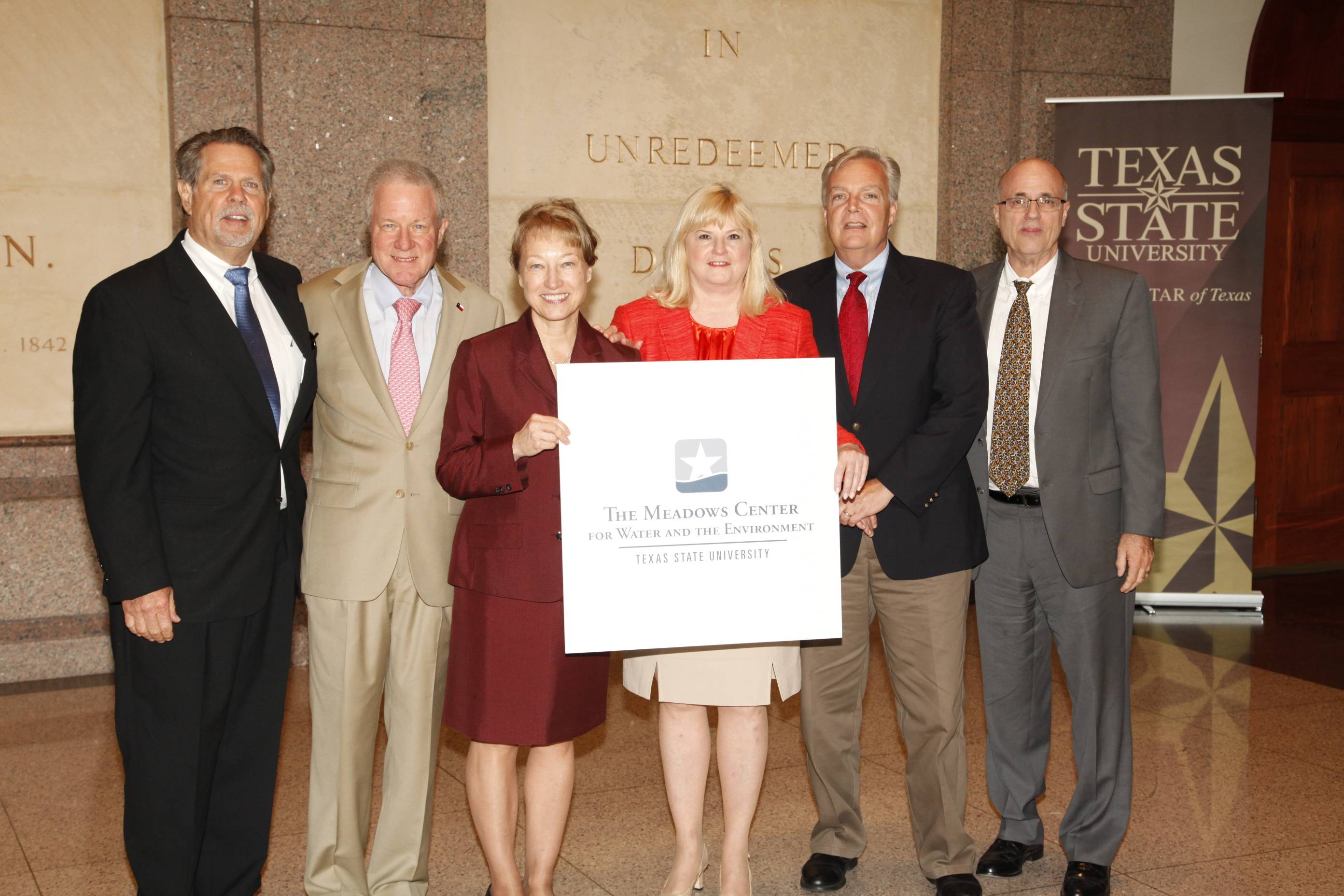 Two smiling women wearing red skirt suits hold up a placard and are flanked on either side by several men in suits. The placard says "The Meadows Center for Water and the Environment -- Texas State University".