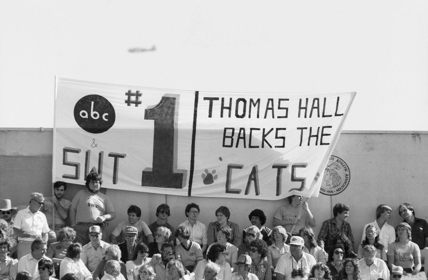 An aged black and white photo of a crowd of people at a sporting event with two men holding a large fabric sign that reads "ABC & SWT #1. Thomas Hall Backs the 'Cats"