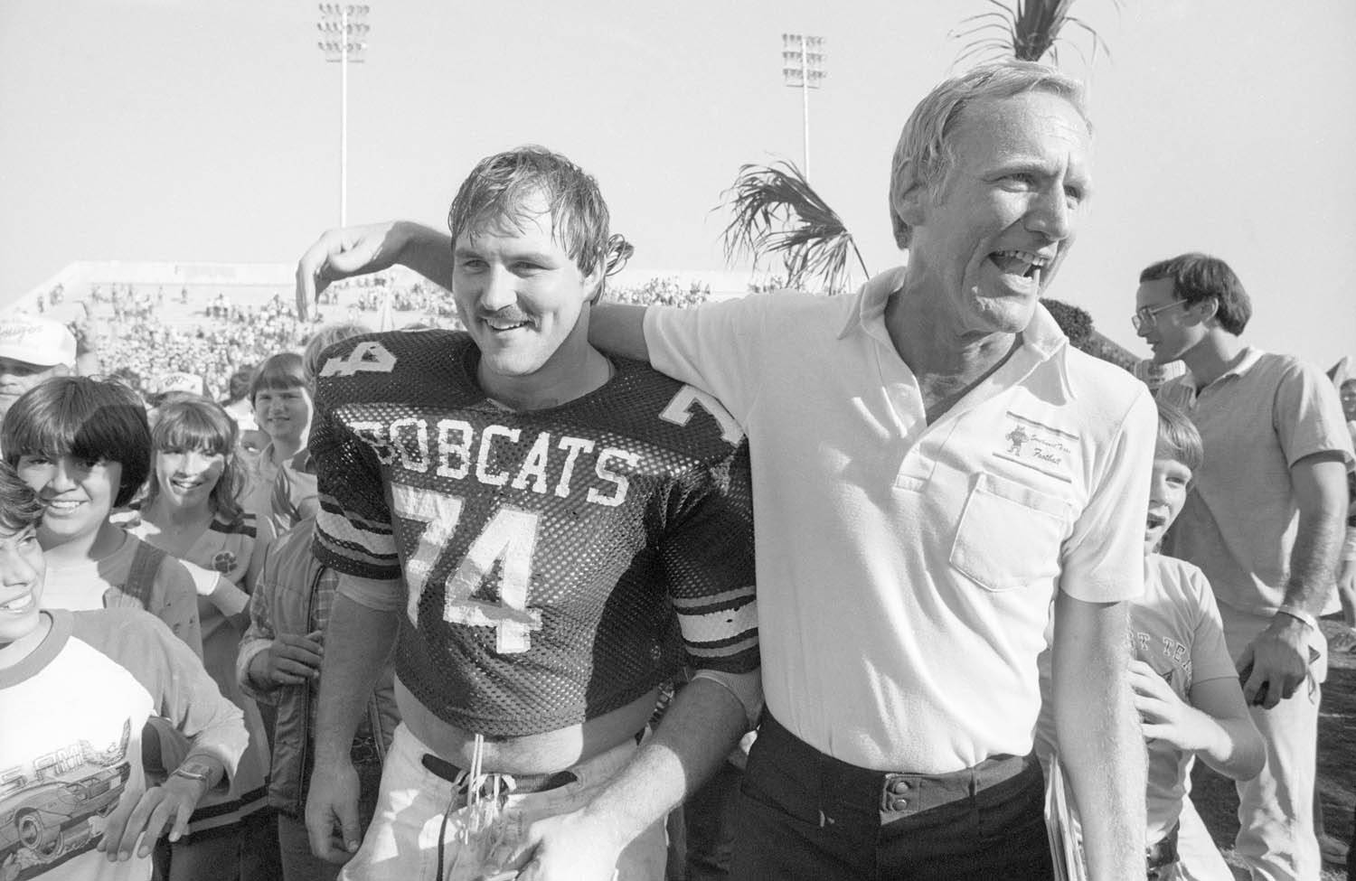 An aged black and white photo of an older male football coach with his arm around the shoulders of a football player wearing a jersey that reads "Bobcats 74". Younger children are in the background looking on in excitement.