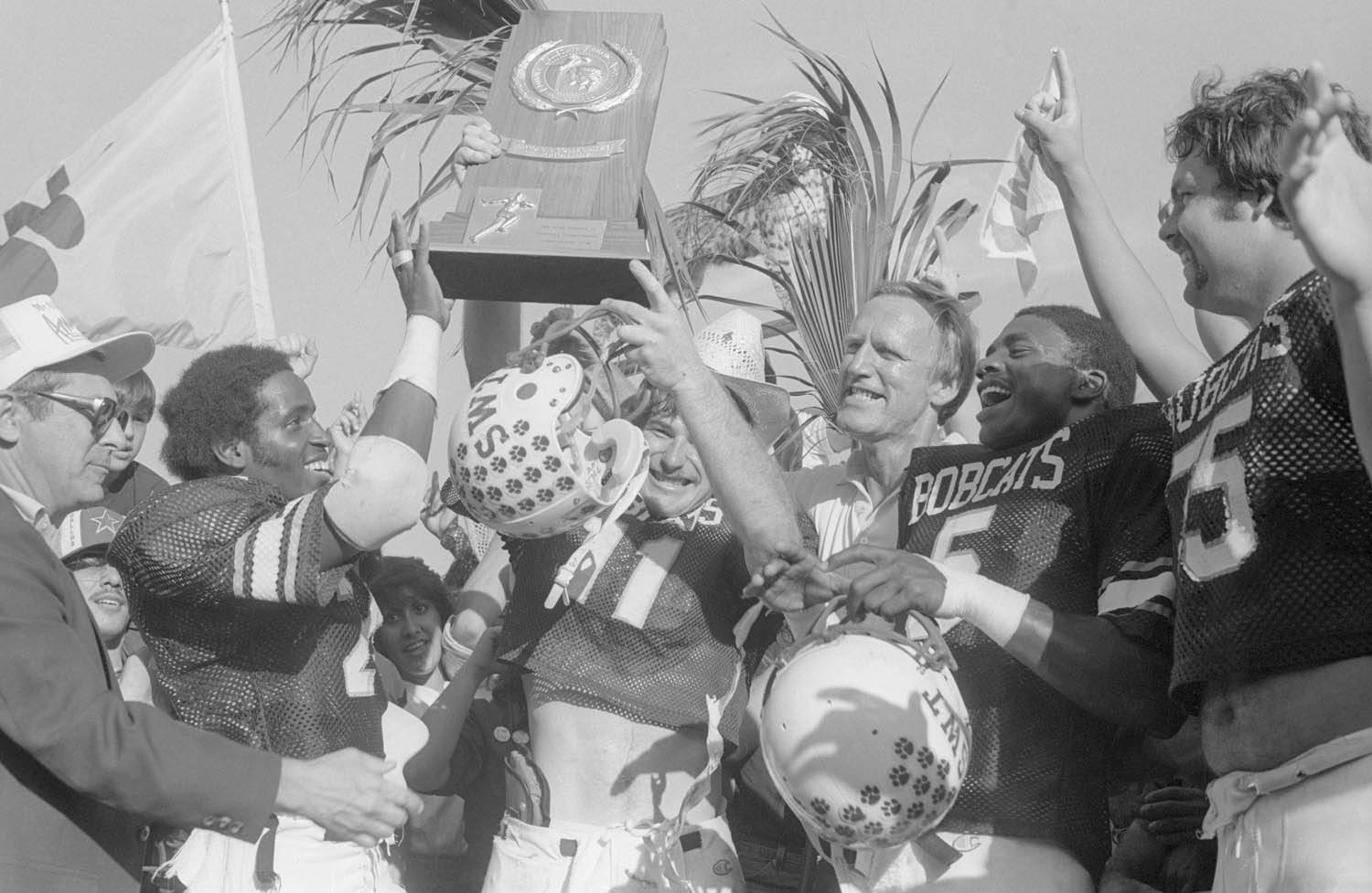 An aged black and white photo of a football player holding up a large trophy, while other players and coaches gather around in celebration.