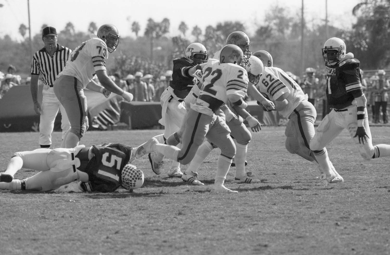 An aged black and white photo of multiple football players in action with an observant referee in the background.