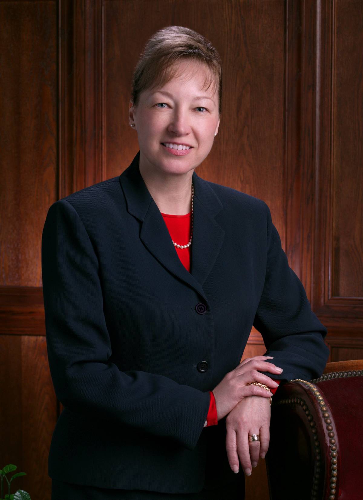 Denise M. Trauth’s presidential portrait. She is wearing a dark suit with a red undershirt. She appears jovial with a smile on her face. 